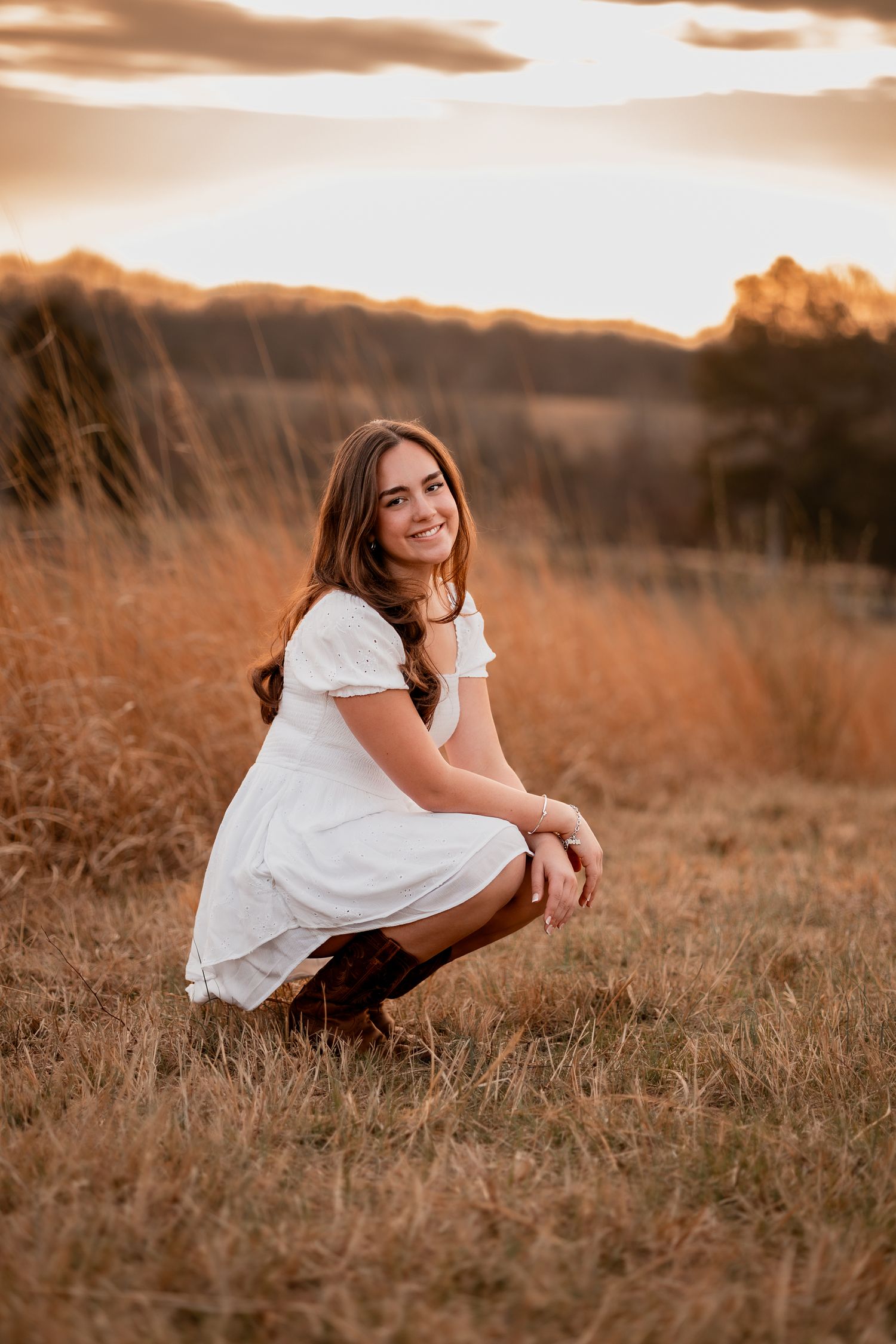 Young woman in white dress and boots crouching in golden field at sunset, smiling warmly at camera.