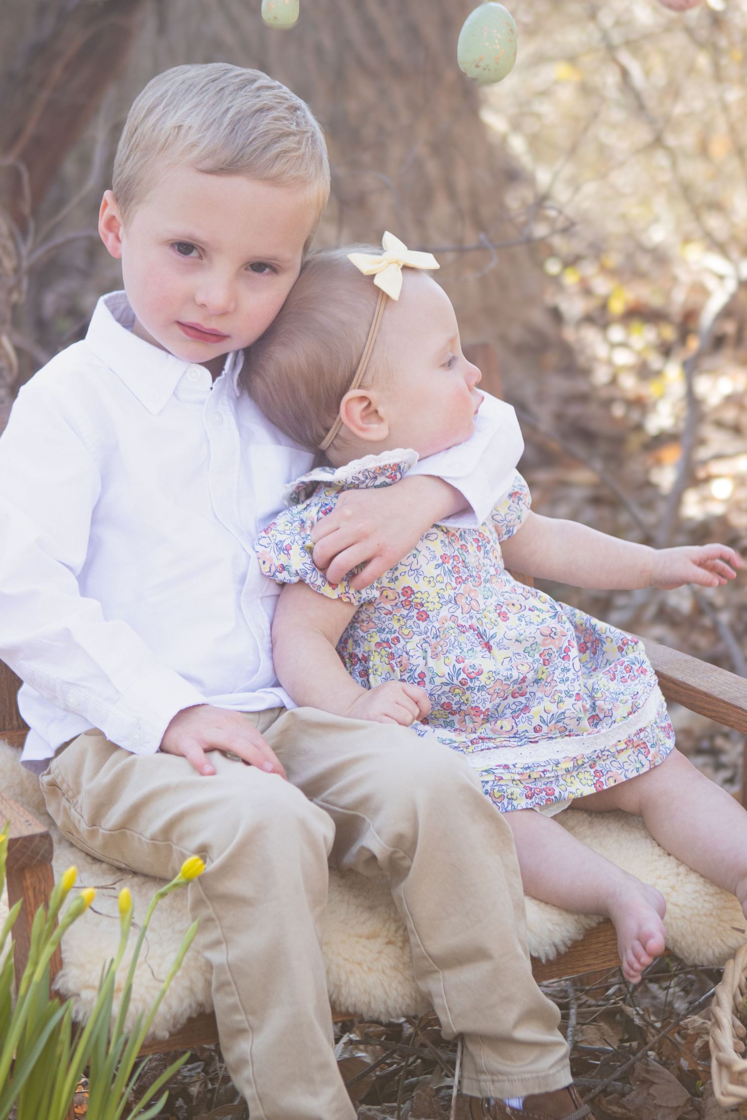 Two siblings share a tender moment on a wooden bench surrounded by spring decor.
