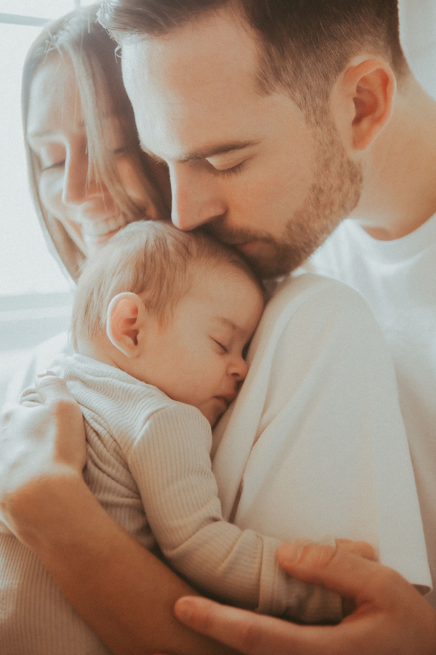 Intimate family moment captured in soft light showing tender embrace between parent and infant.