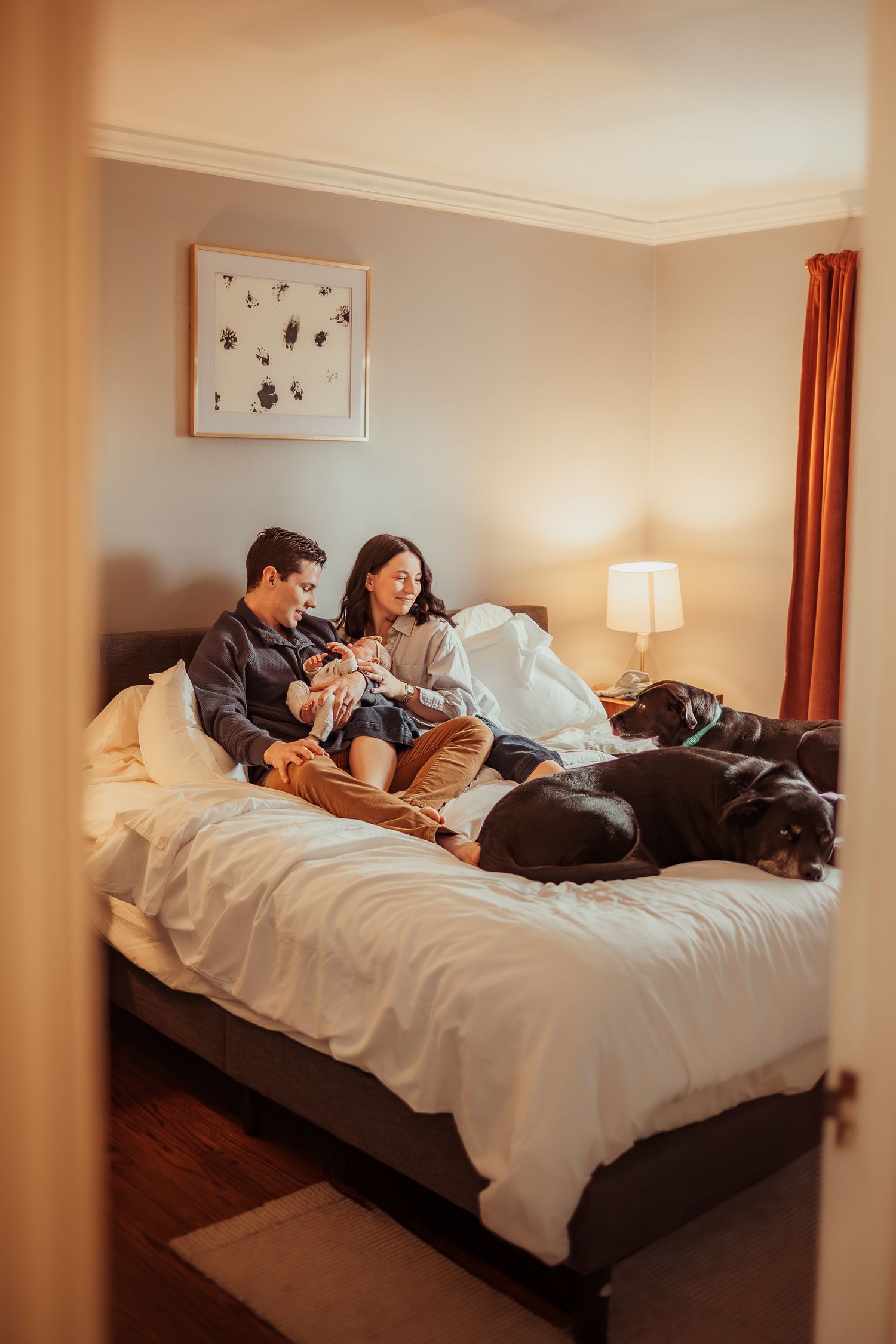 A family relaxing together on a white bed in a cozy bedroom with orange curtains and wall decorations.