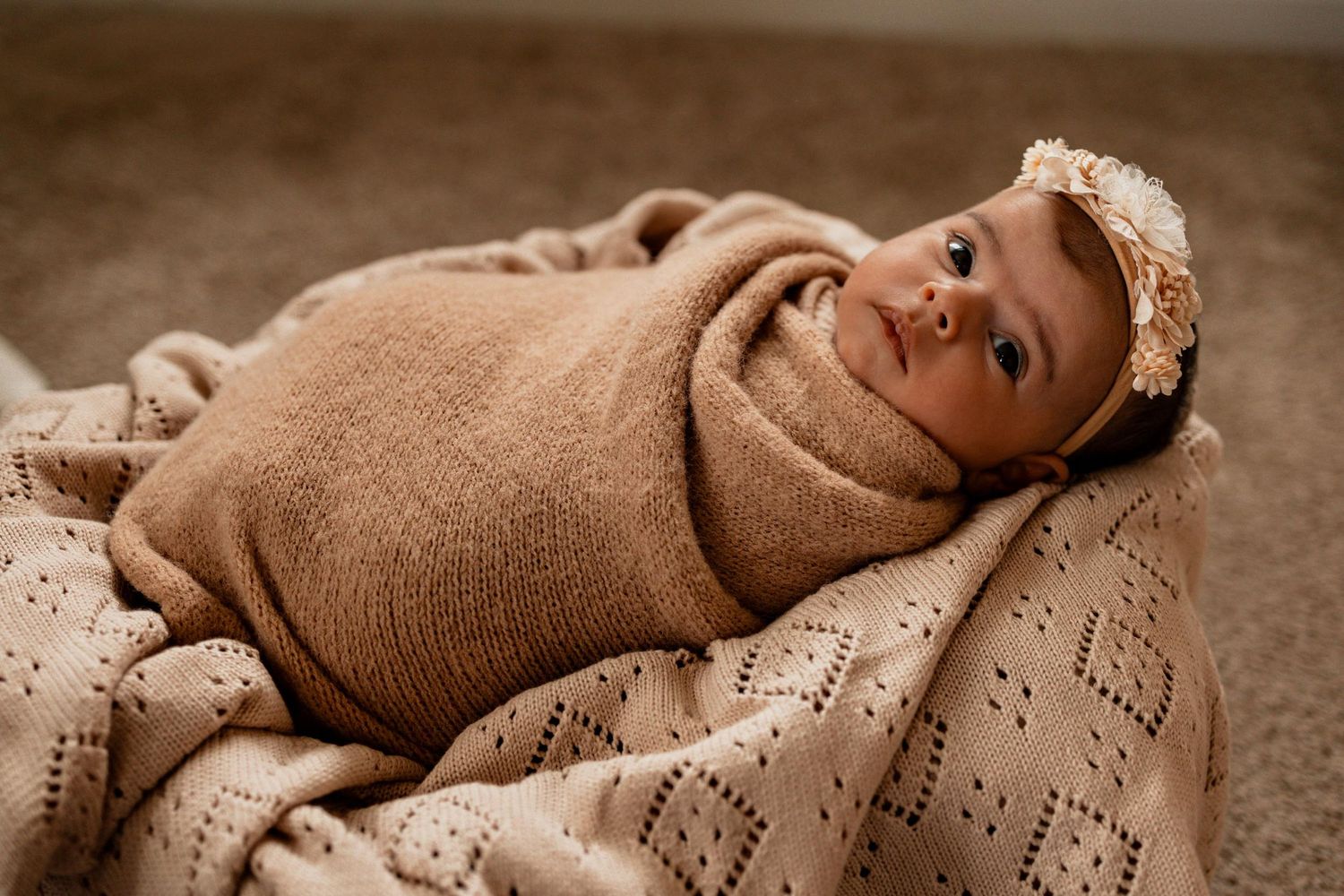 Newborn baby swaddled in a tan knit wrap, wearing a floral headband, resting peacefully in a cozy blanket nest.