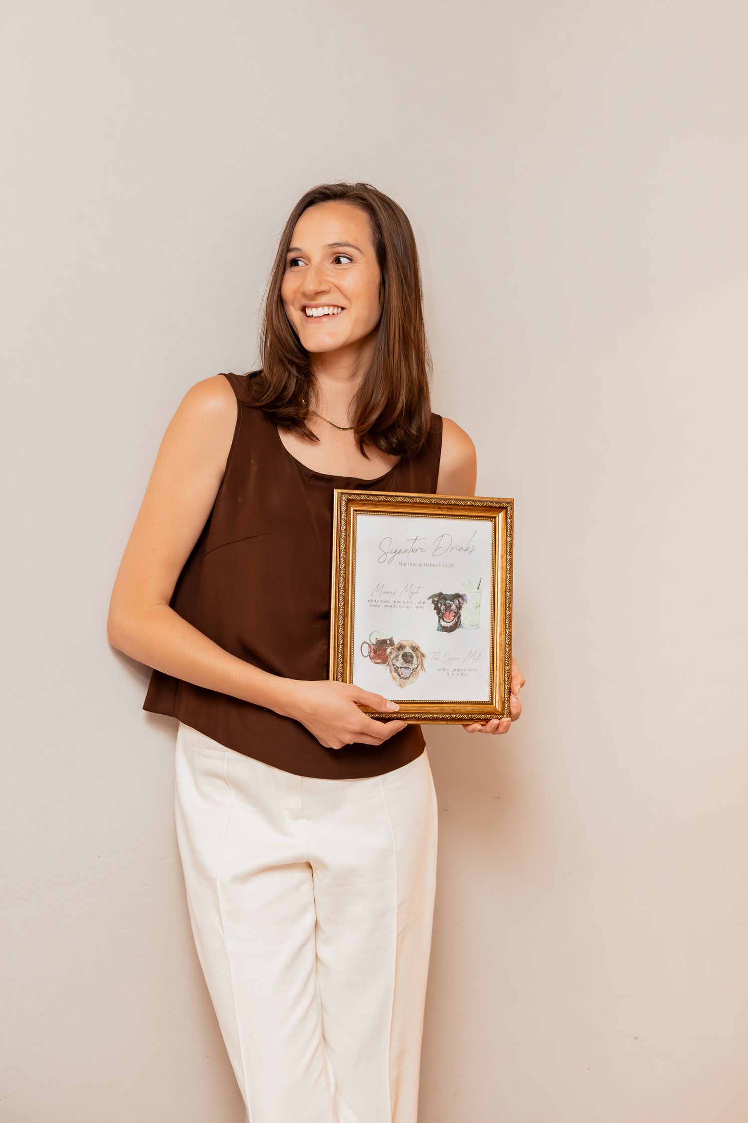Someone wearing a brown tank top and white pants holds a gold-framed artwork of pet portraits against a neutral wall.