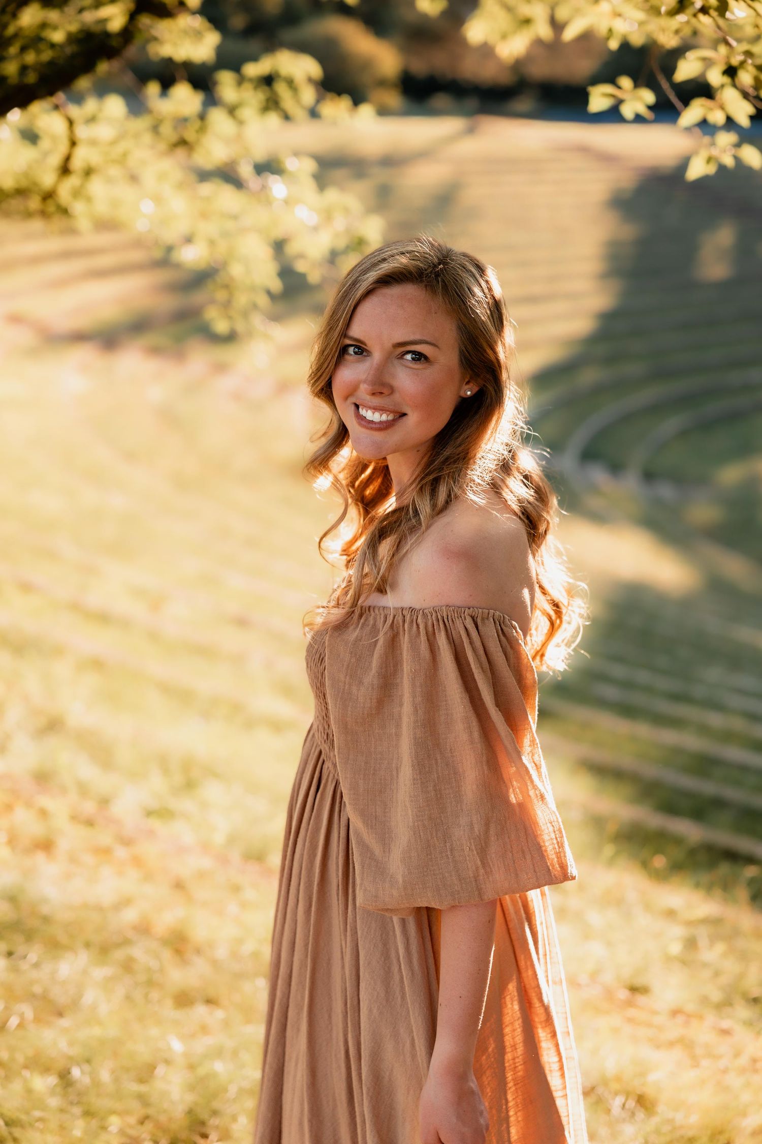 Smiling woman in off-shoulder beige dress standing in sunlit green park with golden hour light.