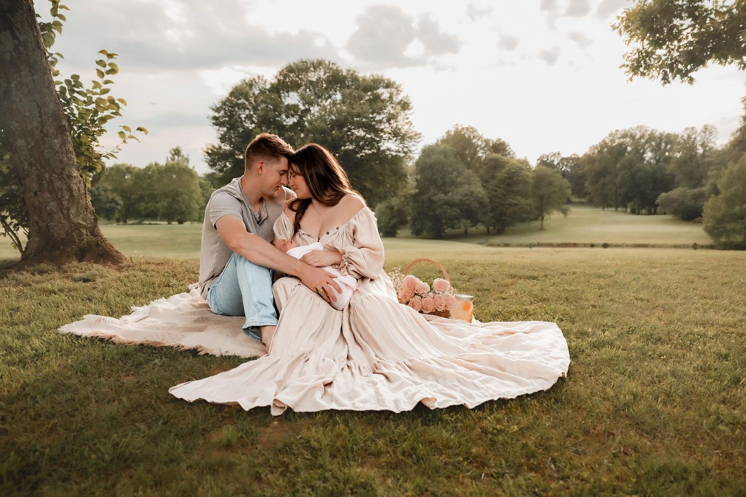 Couple sitting on blanket in lush green park, woman in flowing dress, sharing romantic moment under trees at golden hour.