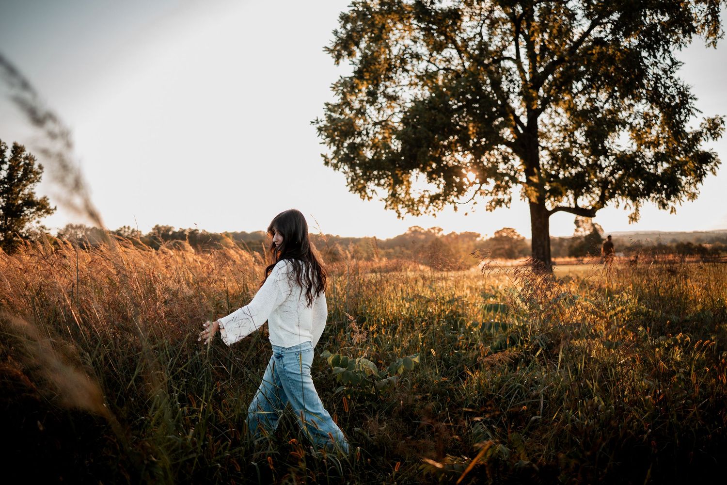 Someone wearing a blue dress twirls in a golden field at sunset as warm light illuminates the scenic landscape.