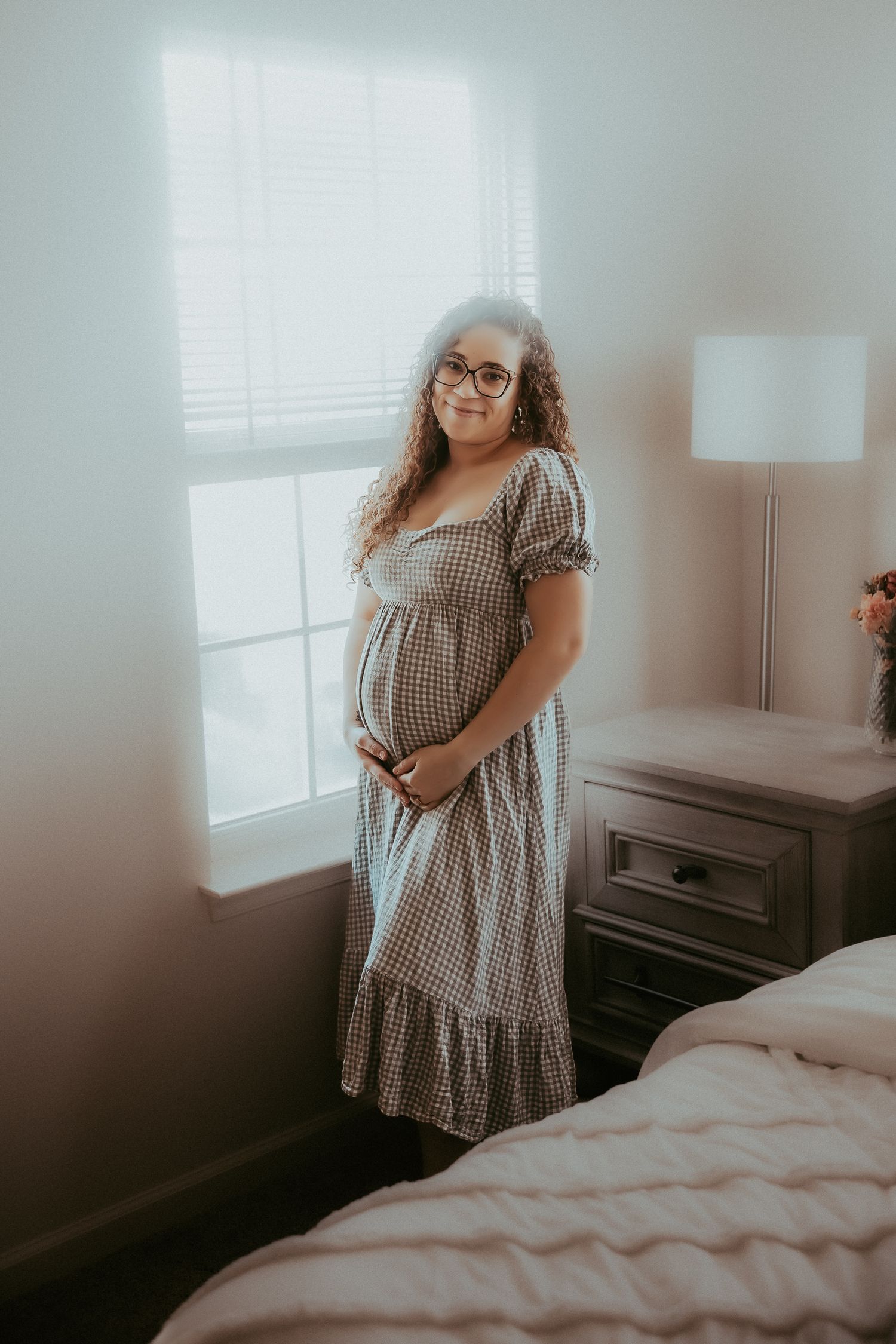 Pregnant woman in gingham dress holding baby bump, standing by bedroom window with curly hair and glasses.
