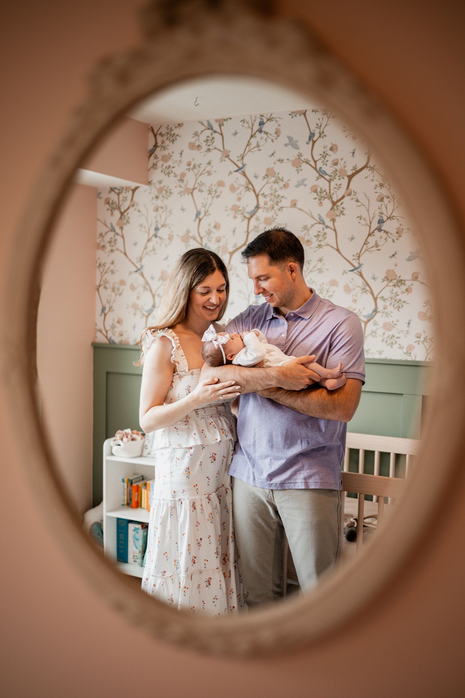 A couple holds their newborn baby while standing in a nursery, viewed through an oval mirror frame.