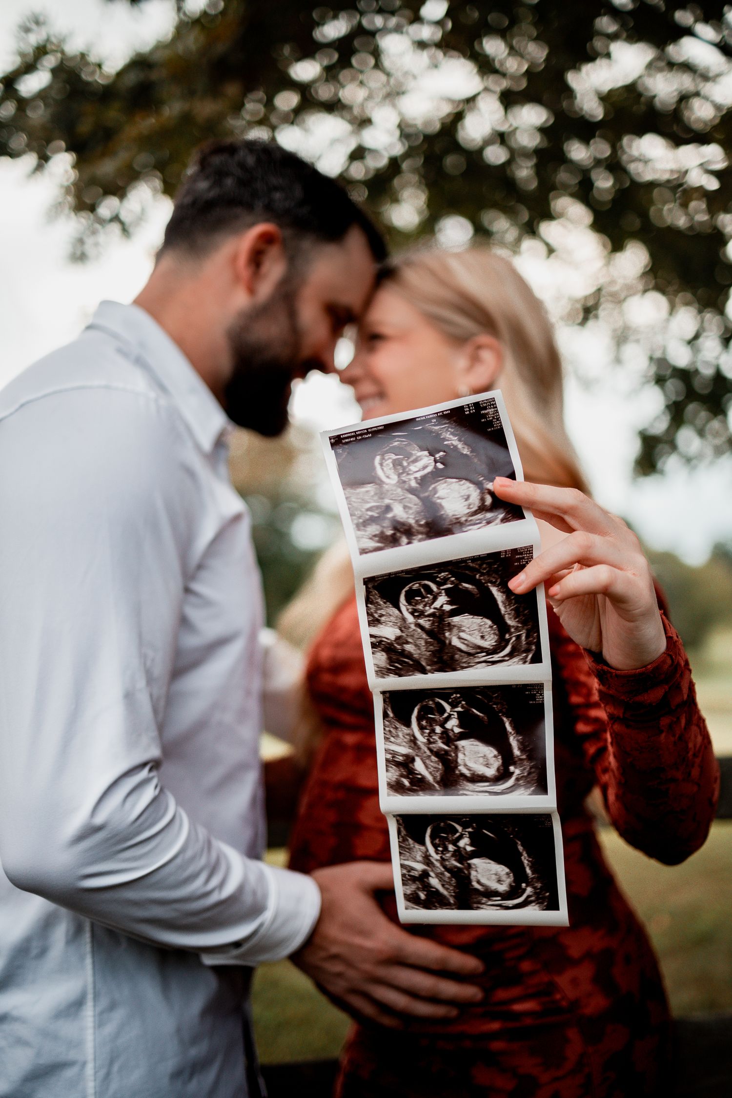 A couple holds ultrasound photos while embracing outdoors in a tender pregnancy announcement moment.