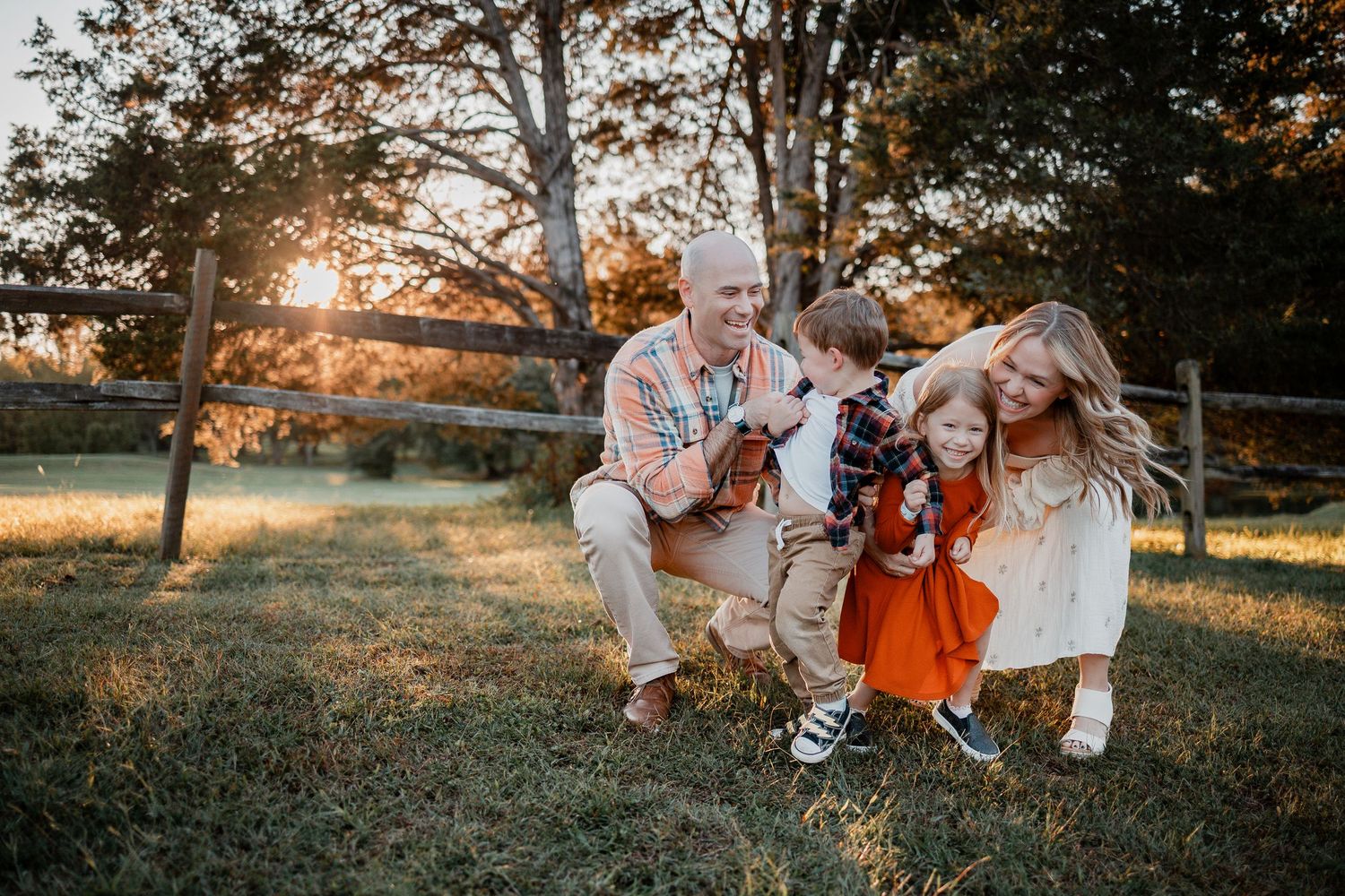 A family in coordinating autumn outfits sits together in a sunlit field during golden hour for a portrait session.
