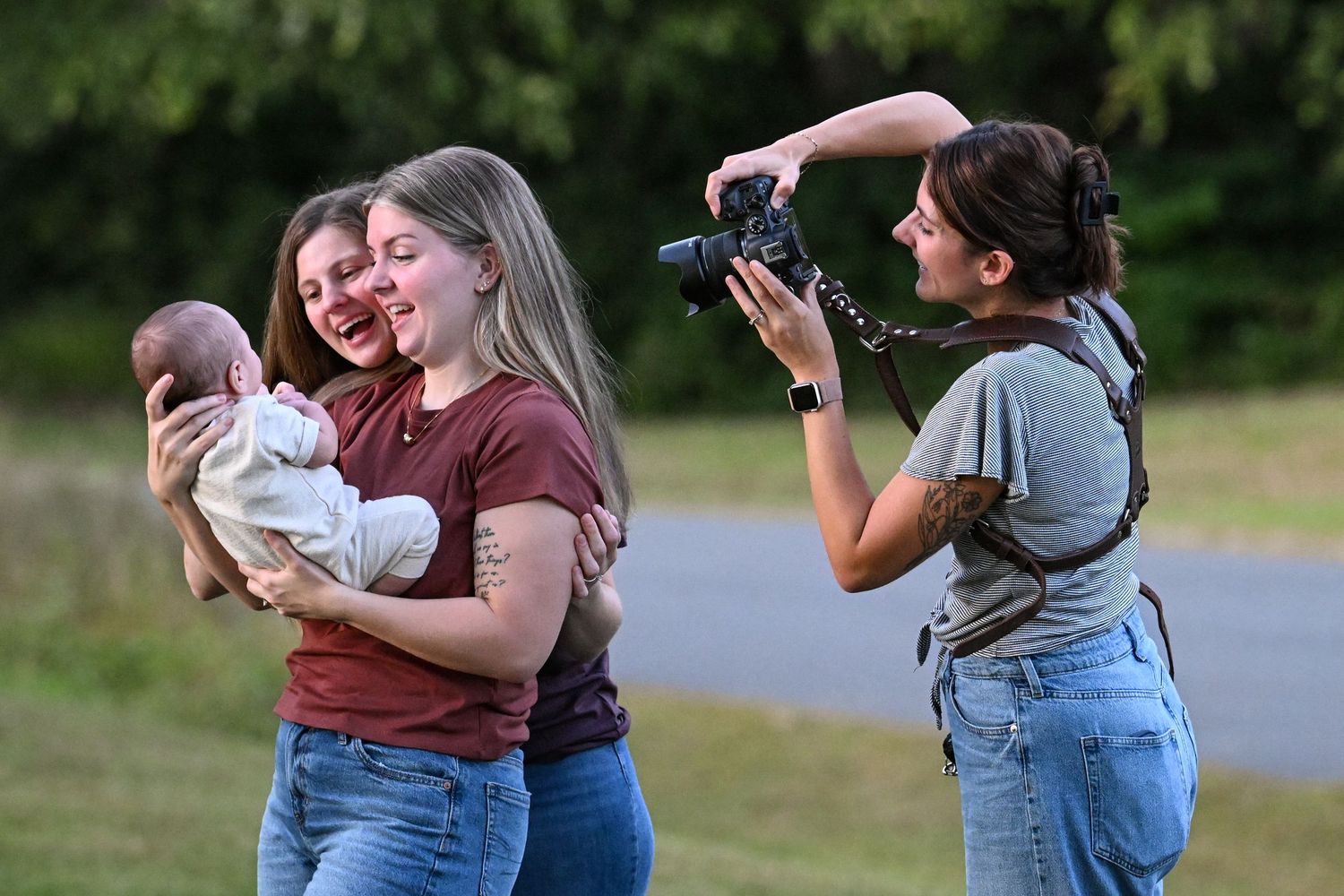 Two women smile holding a newborn baby outdoors while a photographer captures the moment with a camera.