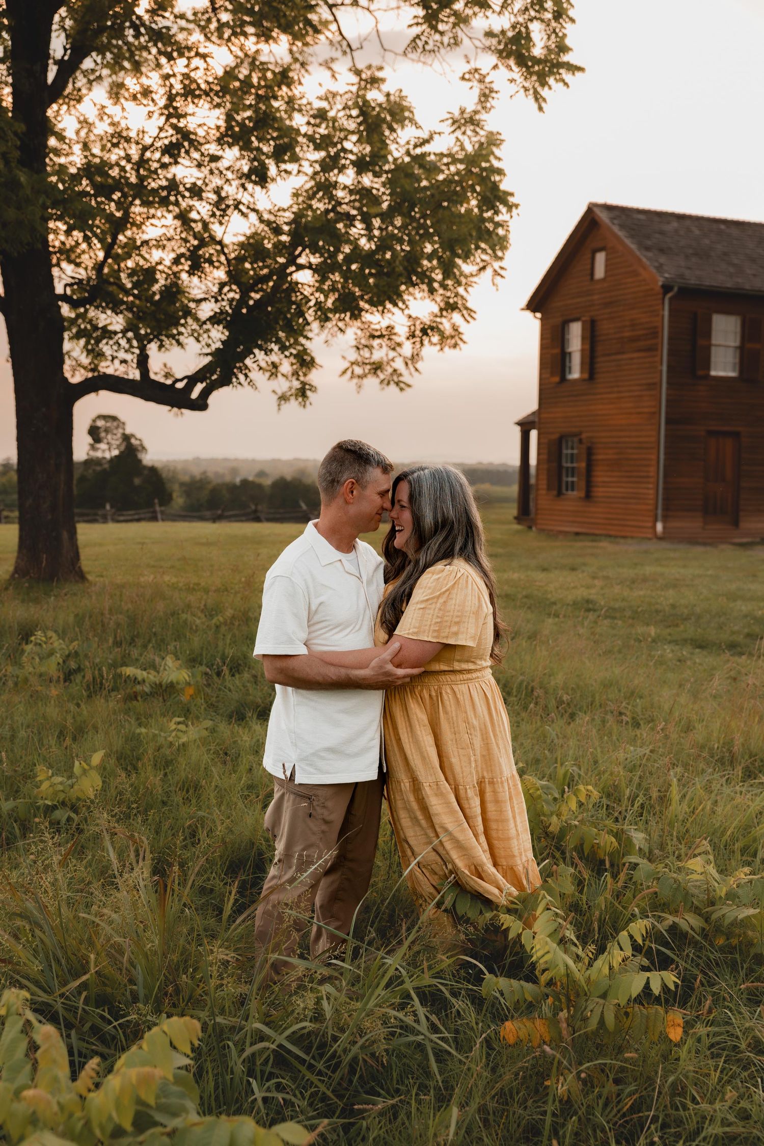 Couple embracing in golden hour light in a green field with a rustic wooden farmhouse and large tree behind them.