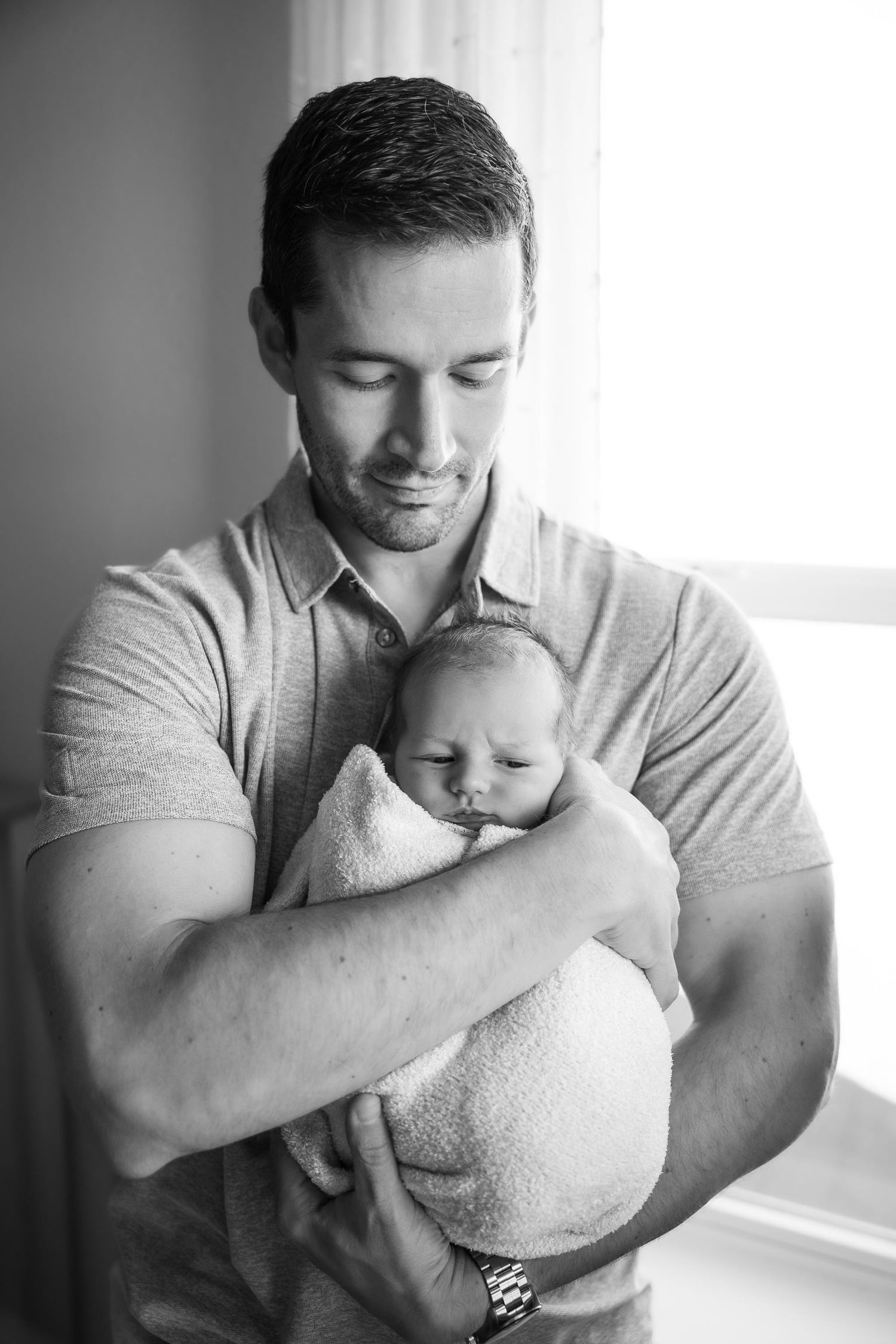 Black and white portrait of father tenderly holding swaddled sleeping newborn baby near a bright window.