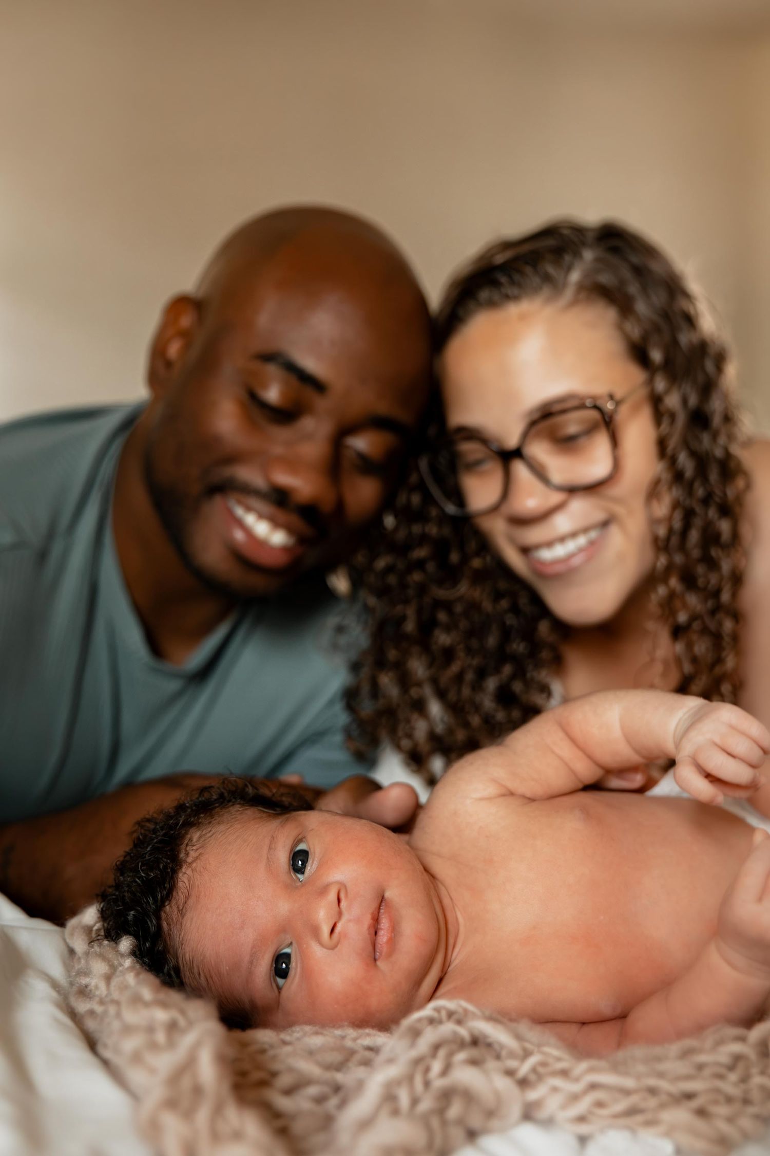 A smiling couple lovingly gazes at their newborn baby lying on a soft blanket in a warm, intimate family portrait.