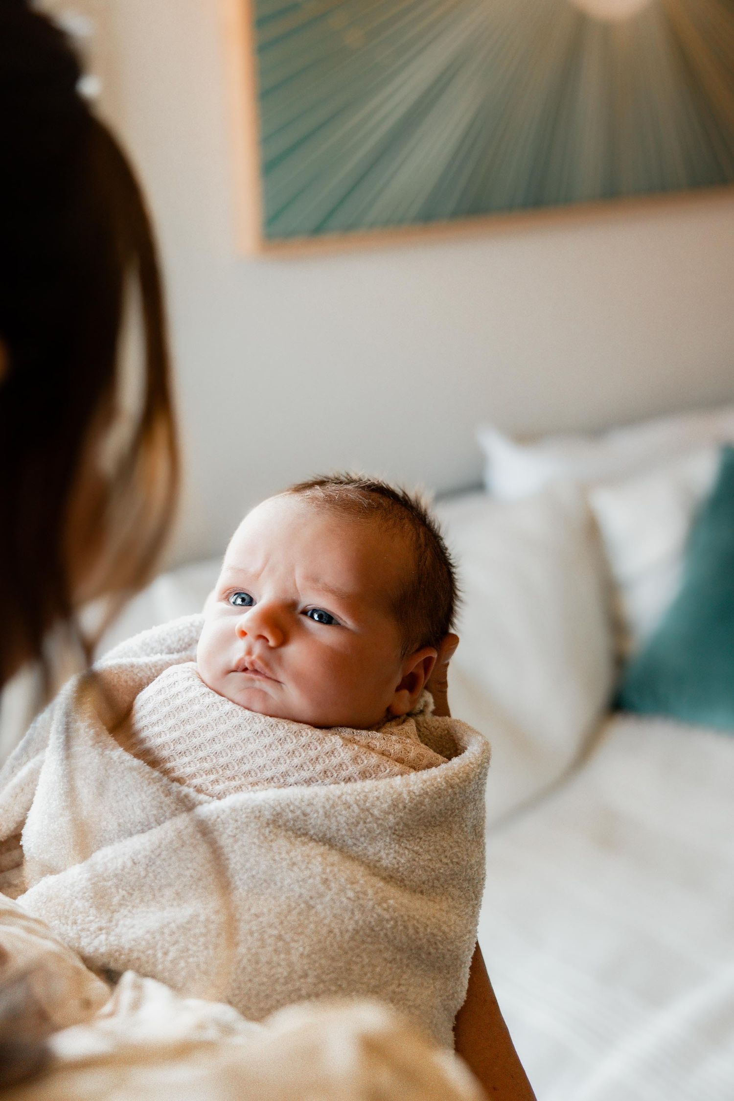 Newborn baby swaddled in a cream blanket with a serious expression, held by a mother in a bright bedroom.