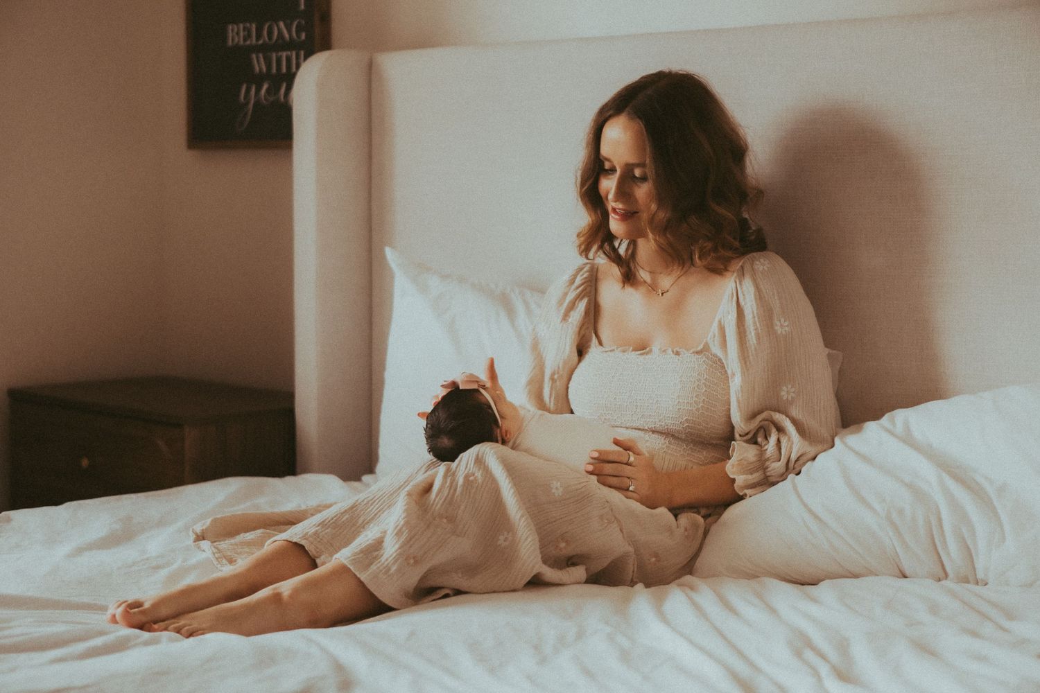 Pregnant woman in white dress sitting on bed cradling her baby bump in a softly lit bedroom.