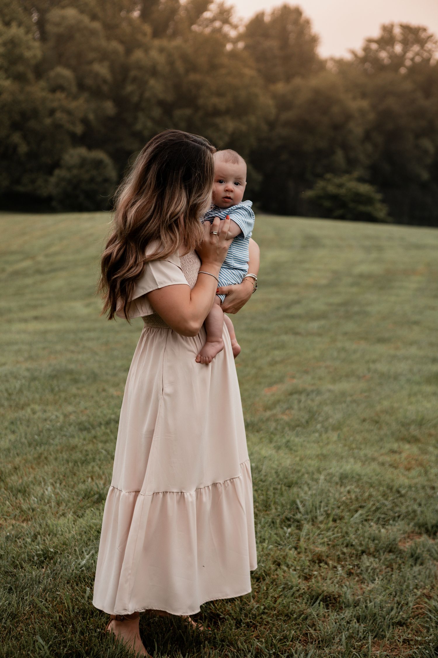 Loving outdoor photoshoot with baby in white dress and blue outfit against green grass background.
