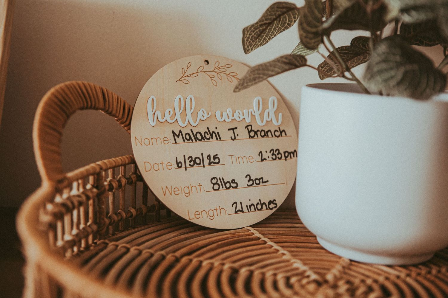 A handwritten birth announcement card on a rattan table with a white potted plant in soft warm lighting.
