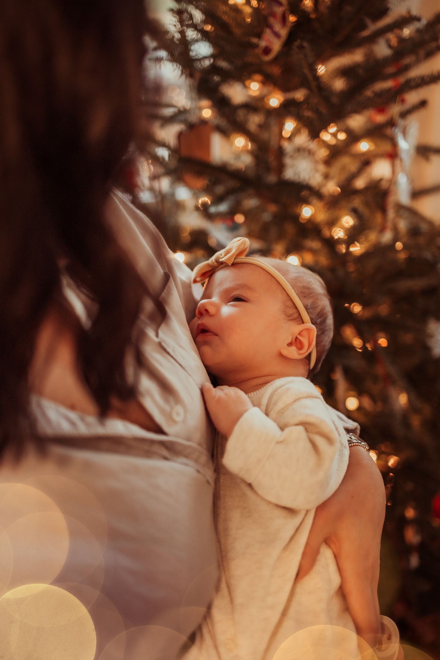 A baby sleeps peacefully in white clothing with twinkling Christmas tree lights glowing in the background.