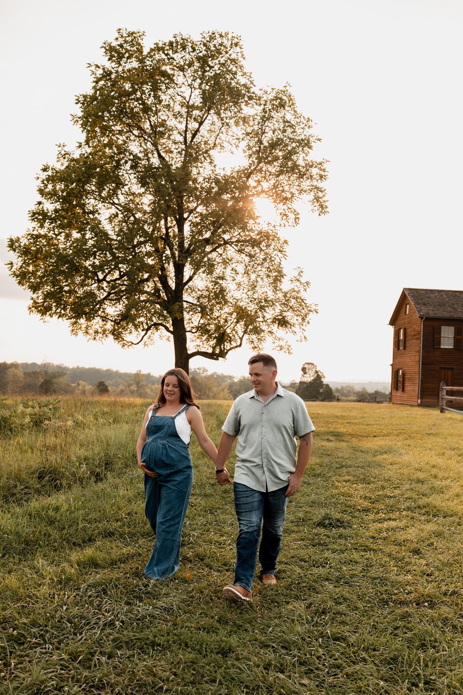 Pregnant couple holding hands walking in golden hour sunlight near a large tree and rustic barn on a green farm.