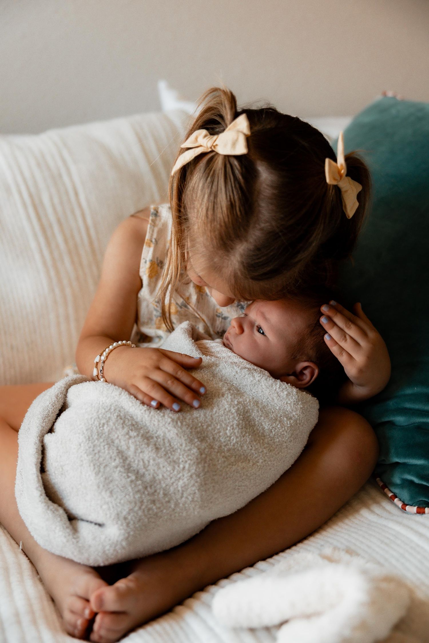 Young girl with bow pigtails gently kissing a swaddled newborn on a couch.