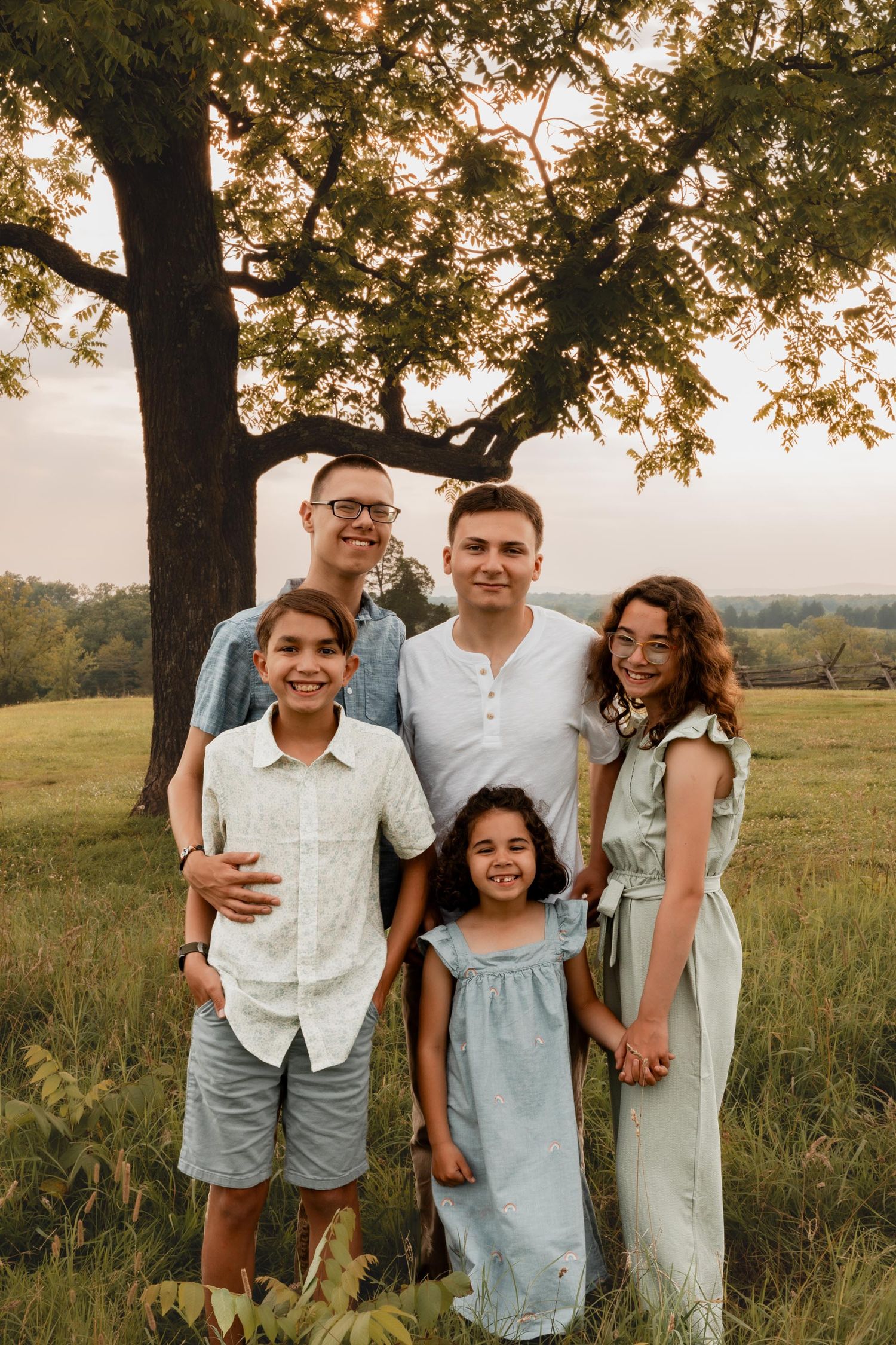 Family of five posing outdoors near a large tree in a green field during golden hour sunset.