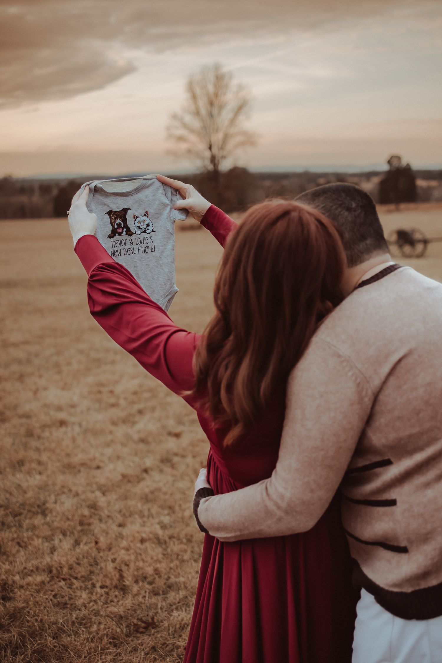 A couple in burgundy dress and beige sweater embrace in a sepia-toned field during sunset.