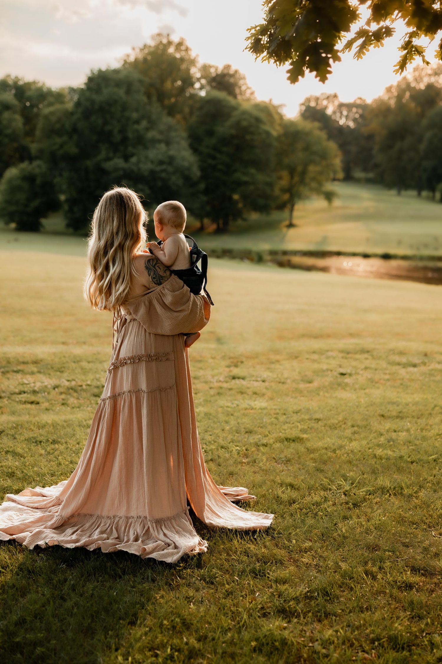 A mother in a flowing peach gown holds her baby while standing in a sunlit green park at golden hour.