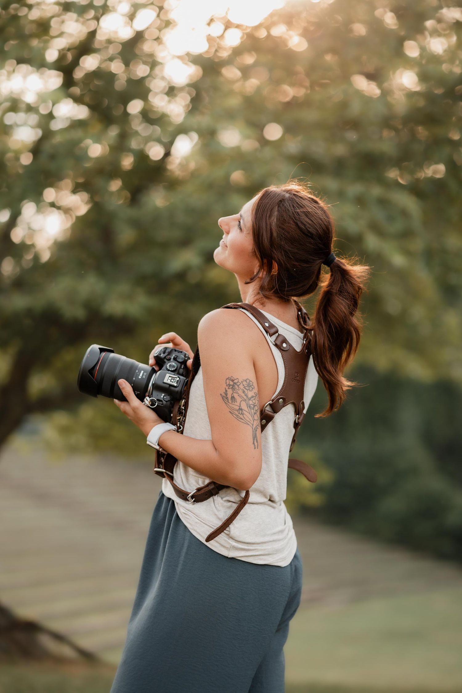 Woman photographer holding DSLR camera outdoors, smiling upward with floral arm tattoo, wearing white tank top at golden hour.