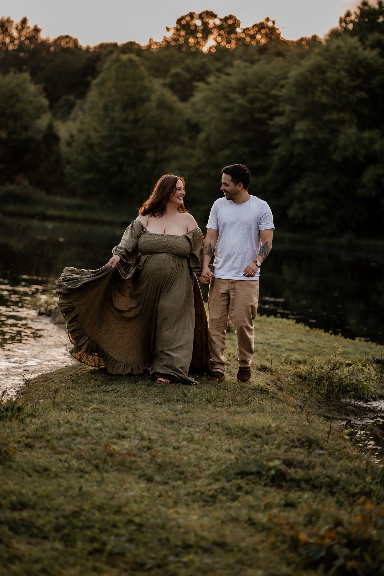 Pregnant couple holding hands and walking by a serene pond at golden hour, woman in flowing olive dress.