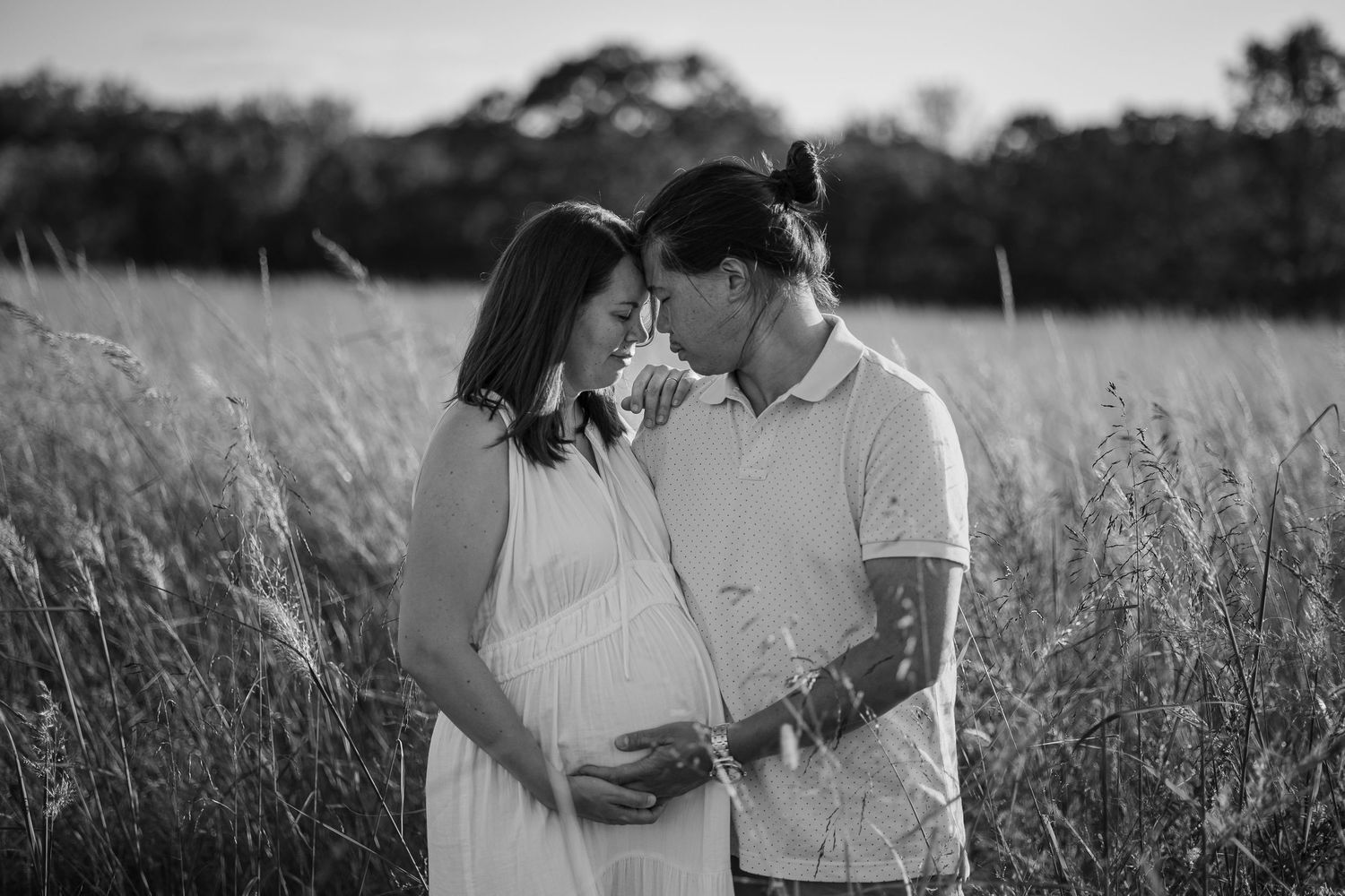 Black and white maternity photo of a couple embracing in a tall grass field at sunset, woman in white dress.