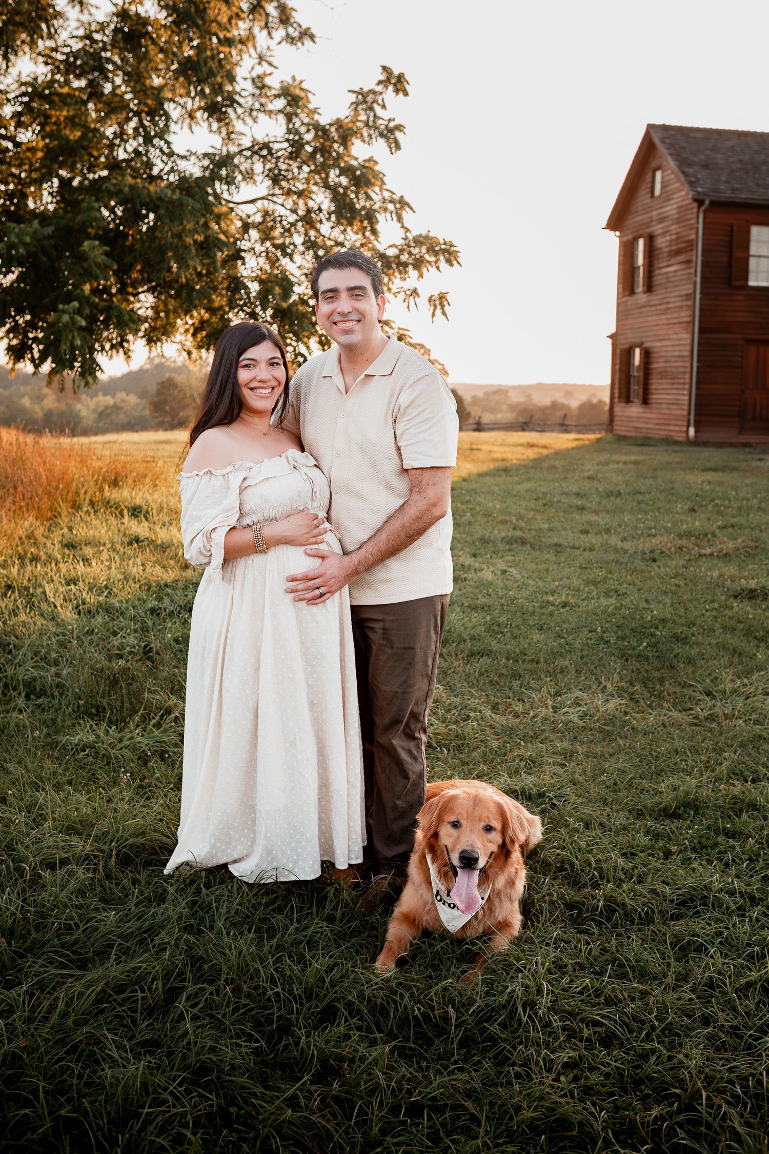 Expectant couple poses with their golden retriever in front of rustic farmhouse at sunset.