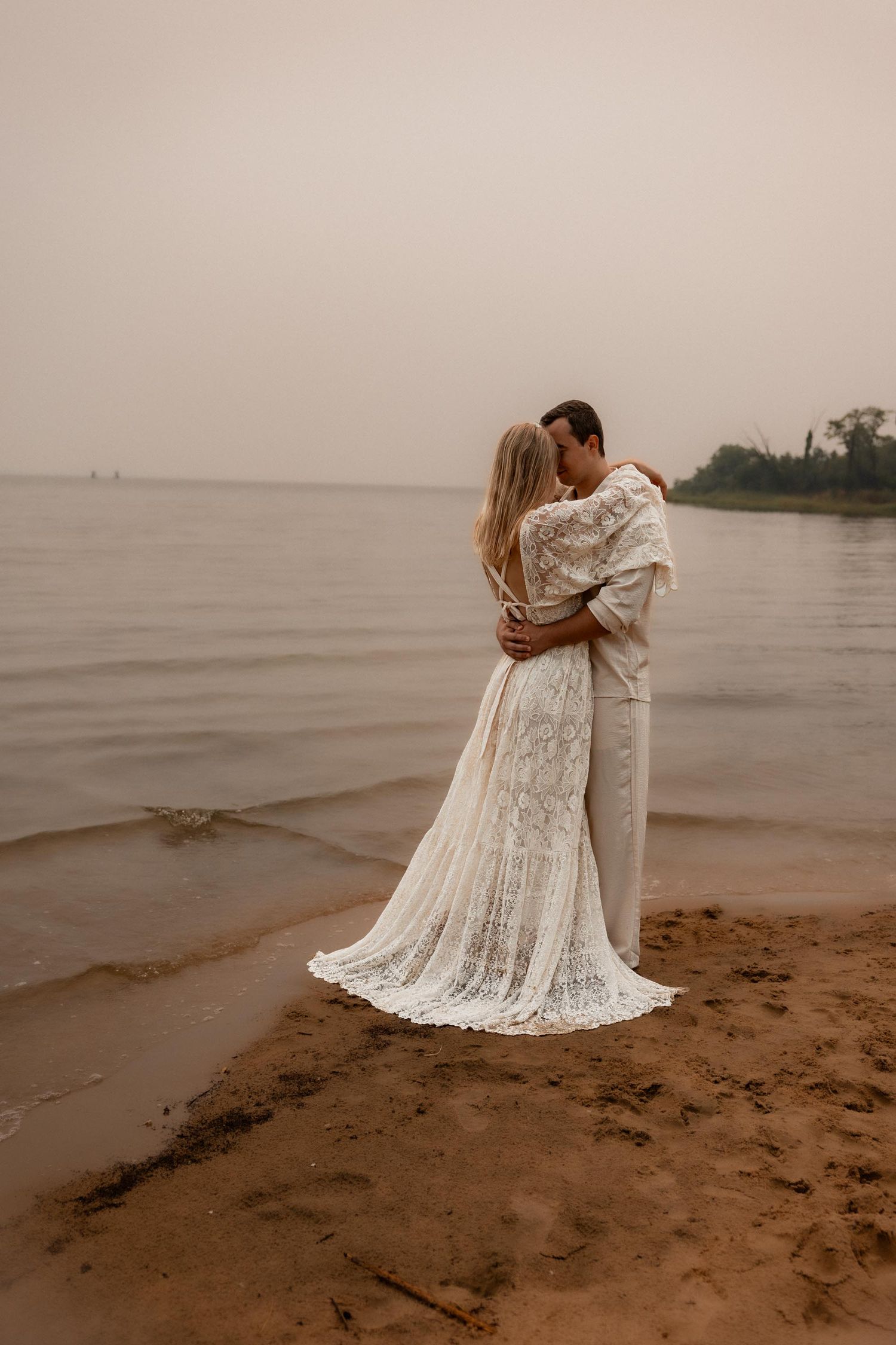 Couple in wedding attire embracing on sandy beach shore with calm water and misty sky backdrop.