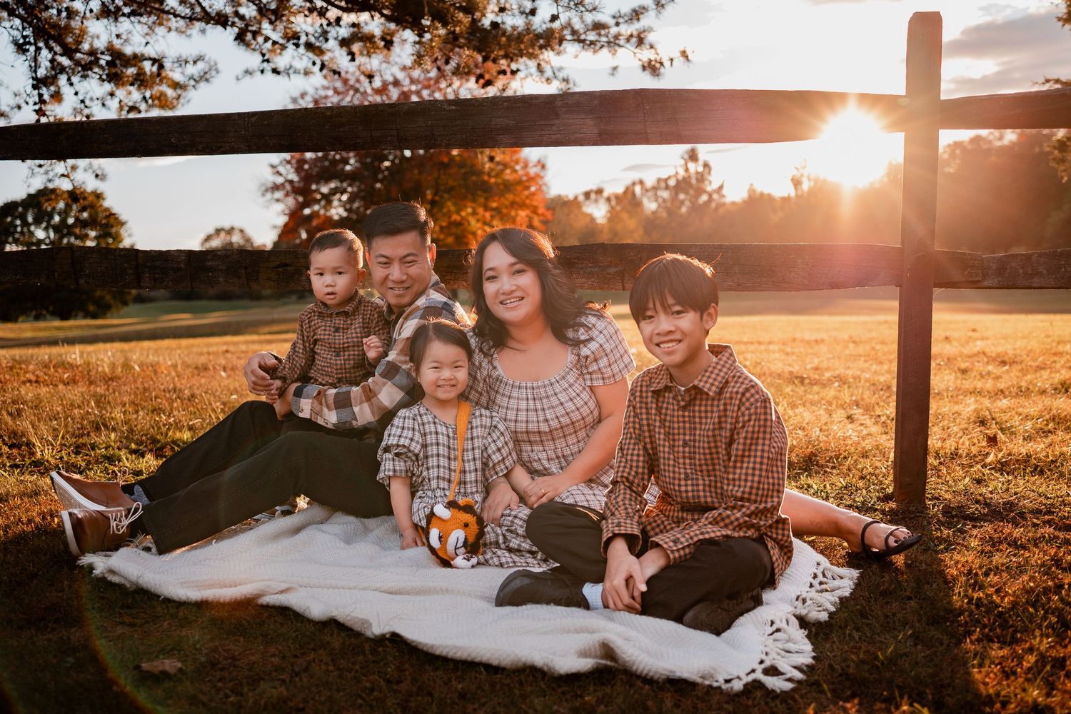 A family relaxes together on a blanket in a golden wheat field at sunset with wooden fence in background.