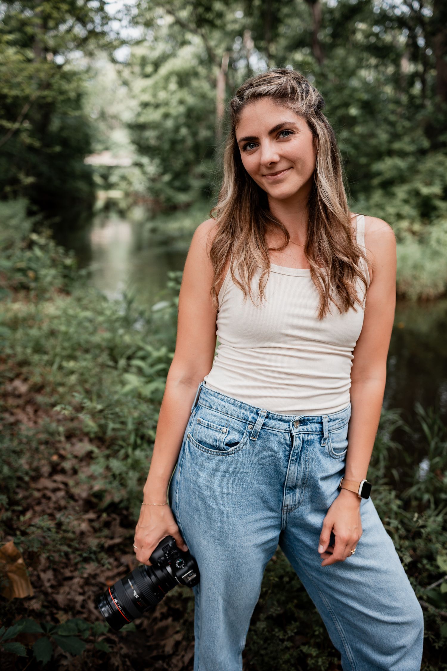 Young woman holding a camera smiling outdoors near a forest stream, wearing jeans and a white tank top.