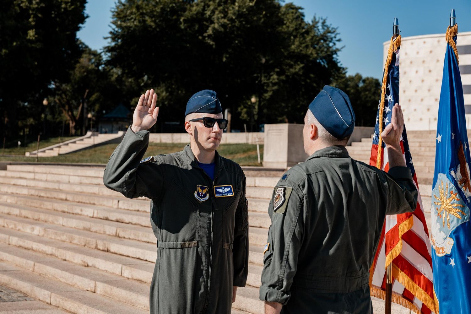 Two Air Force officers in flight suits salute during a military ceremony with American flags outdoors.