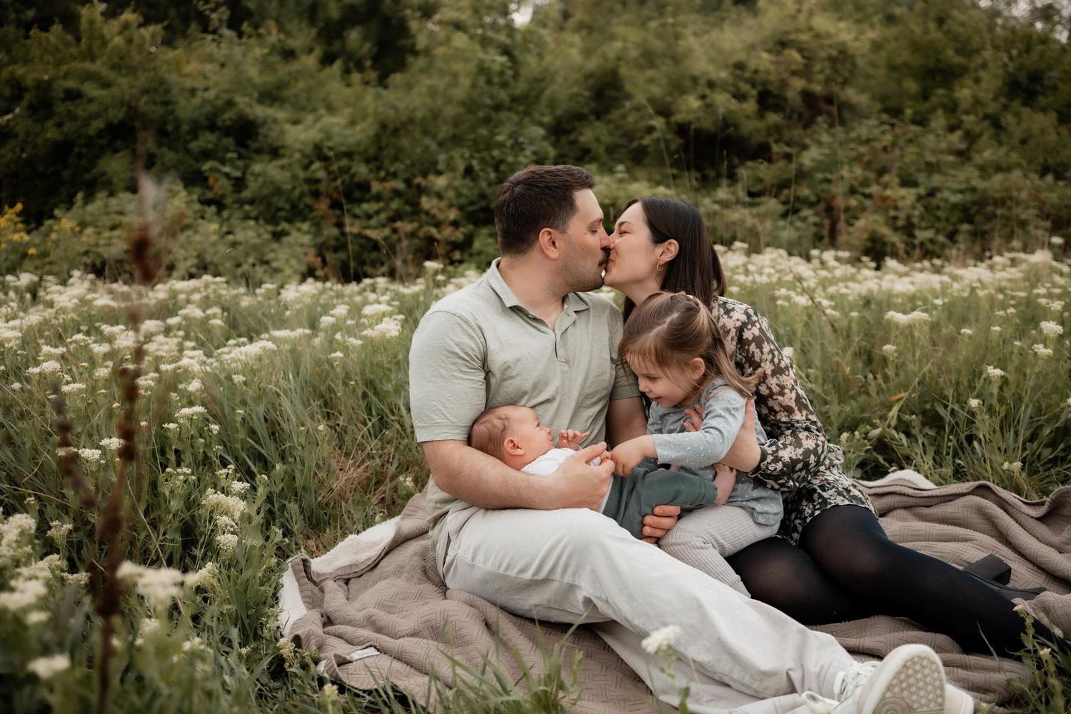 Family of four sitting in a wildflower field, parents kissing while holding newborn and toddler daughter.