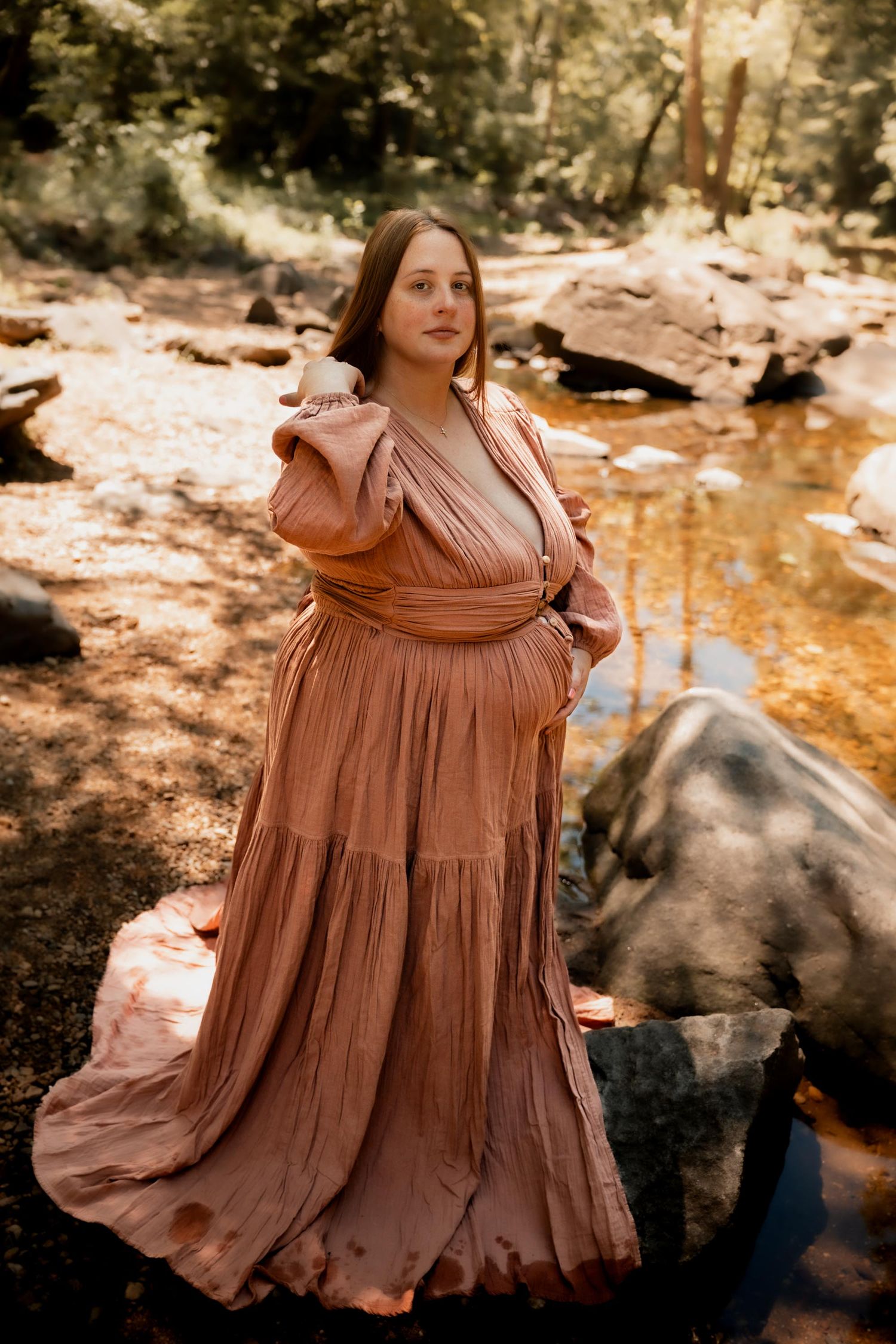 Pregnant woman in flowing terracotta dress standing on rocks by a serene forest creek in natural light.