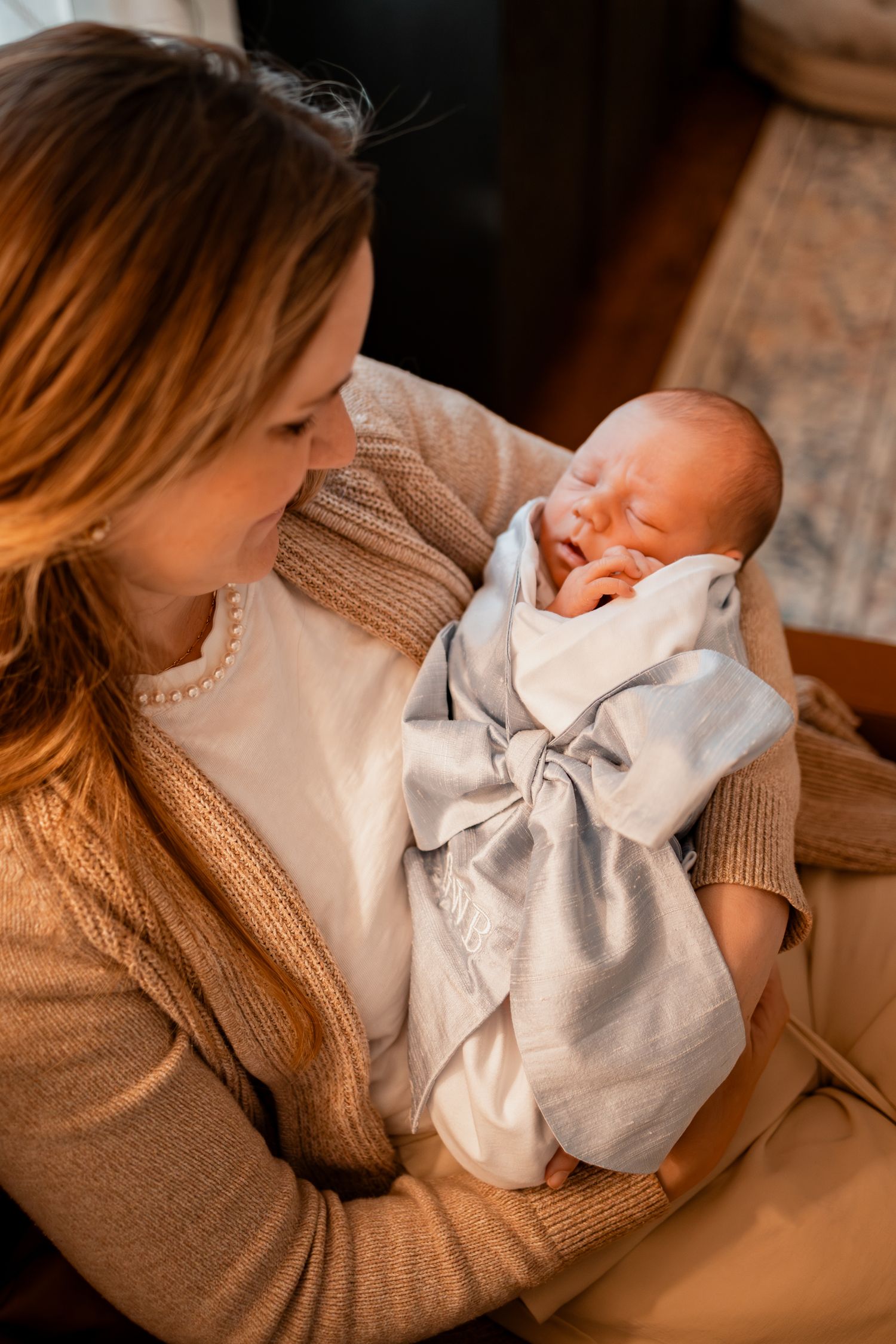 A newborn baby wearing white is cradled in someone's arms wearing a brown knit sweater.