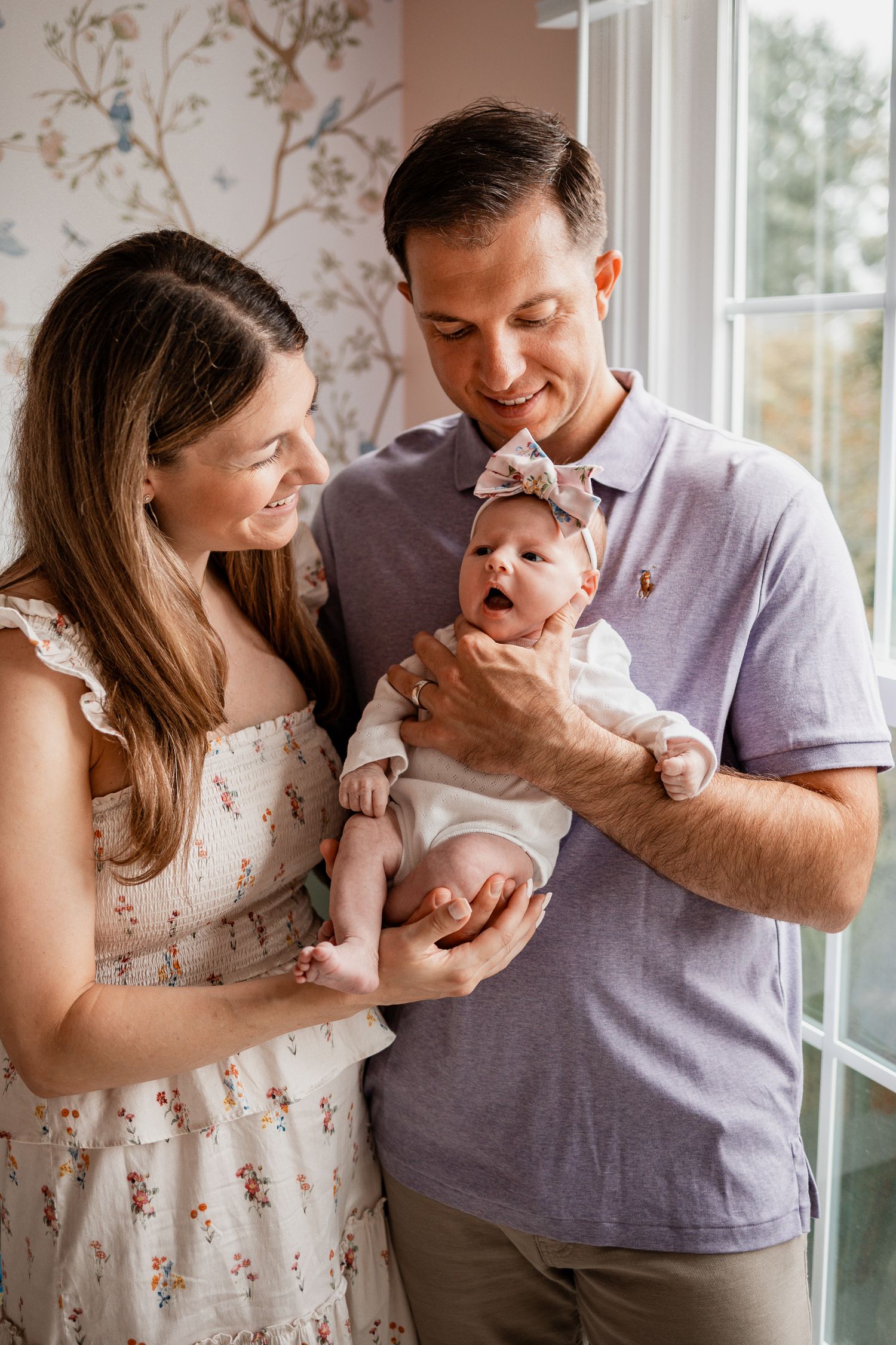 A couple shares a joyful moment with their newborn baby by a window with floral wallpaper in the background.