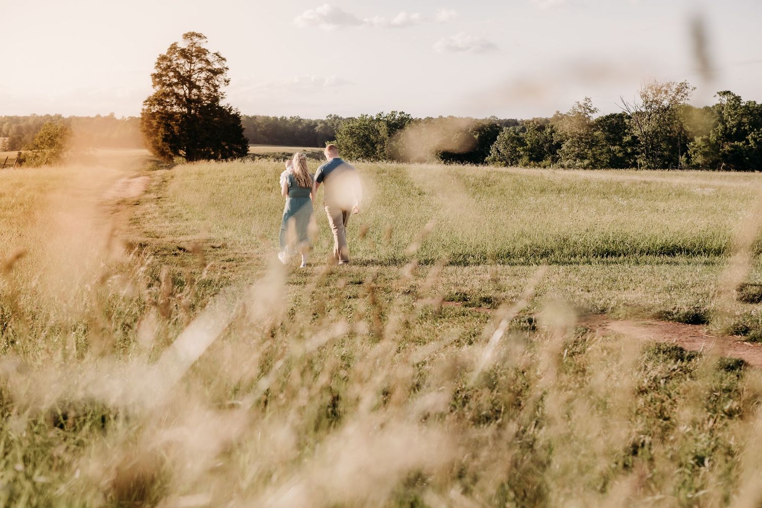 A soft golden-hour scene in a grassy field with someone in a light dress and long hair standing amongst tall grass.