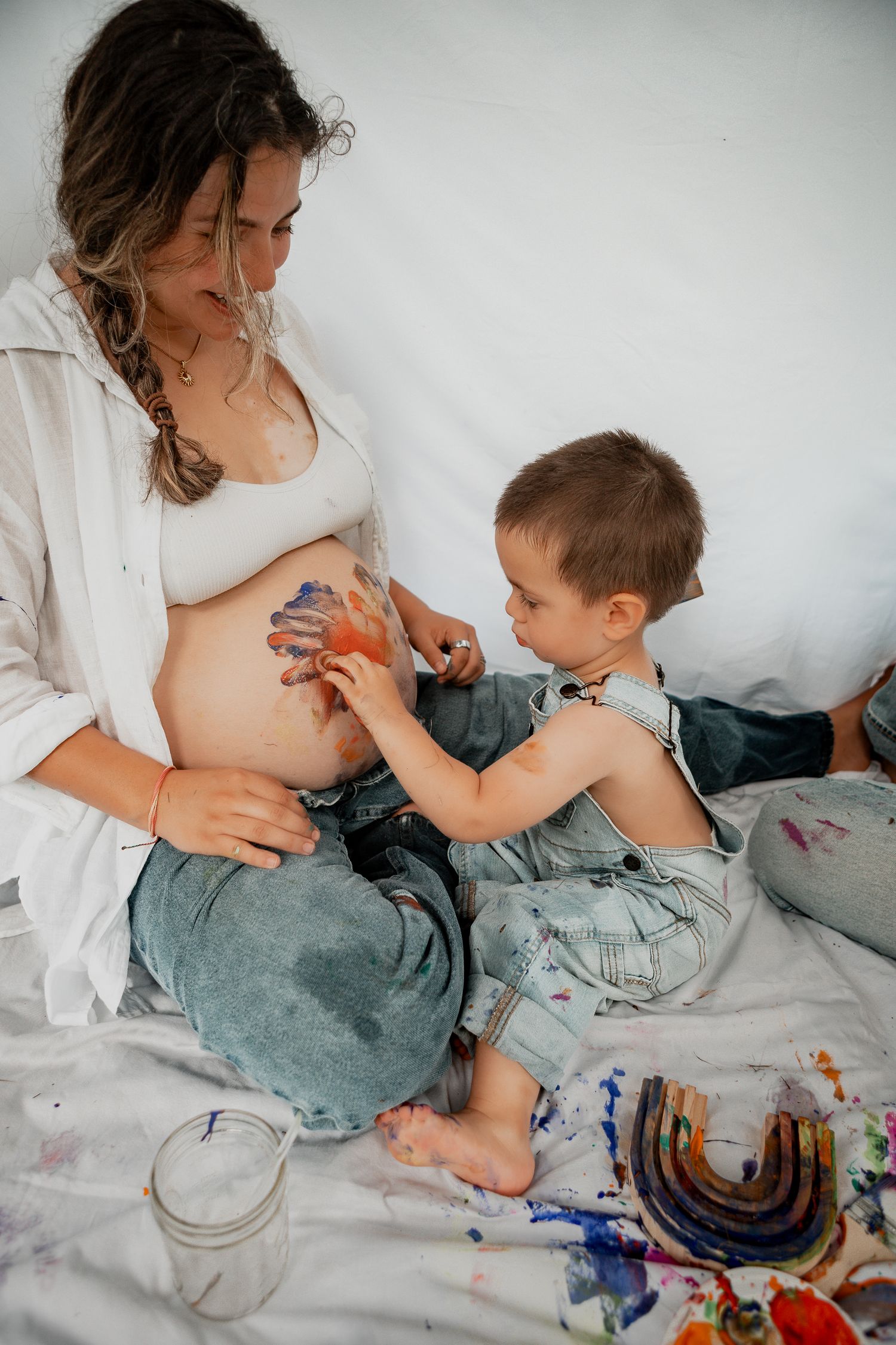 A pregnant woman and young child paint together with colorful paints while sitting on white bedding.
