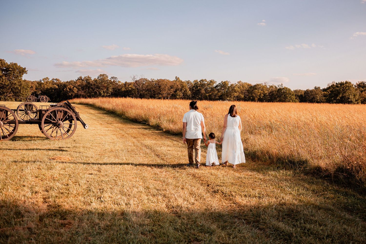 Two people in white walking hand in hand through a golden wheat field at sunset with a vintage bicycle in background.