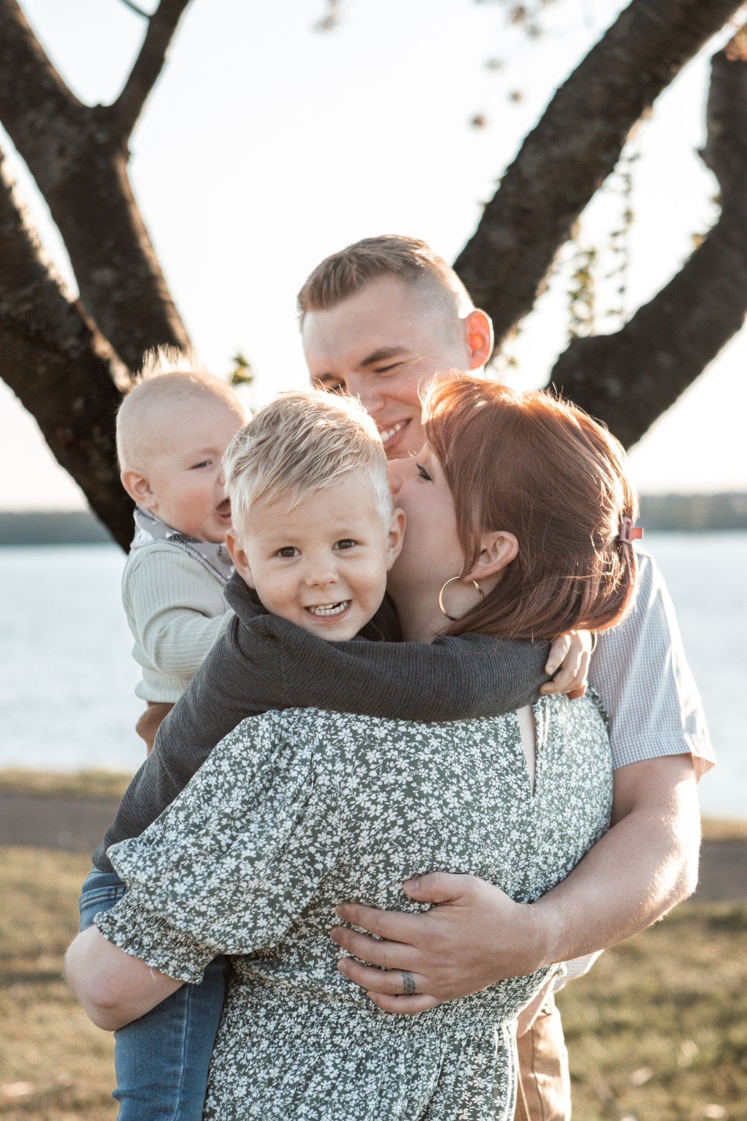 Family sharing an intimate group hug outdoors with a waterfront and tree in the background.