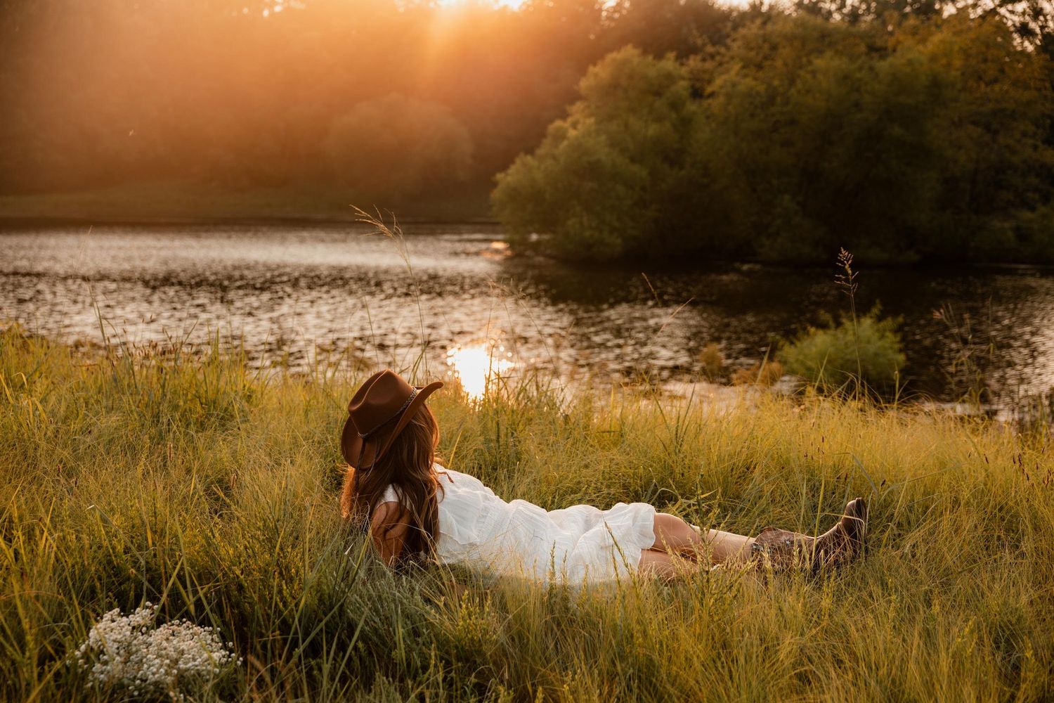 Woman in white dress lying in golden grass by a sunlit lake at sunset surrounded by lush green trees.