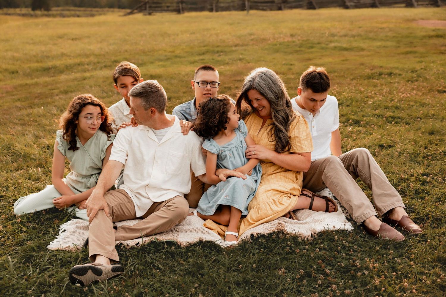 A large family sits together on a blanket in a green field during golden hour, sharing a warm and joyful moment.