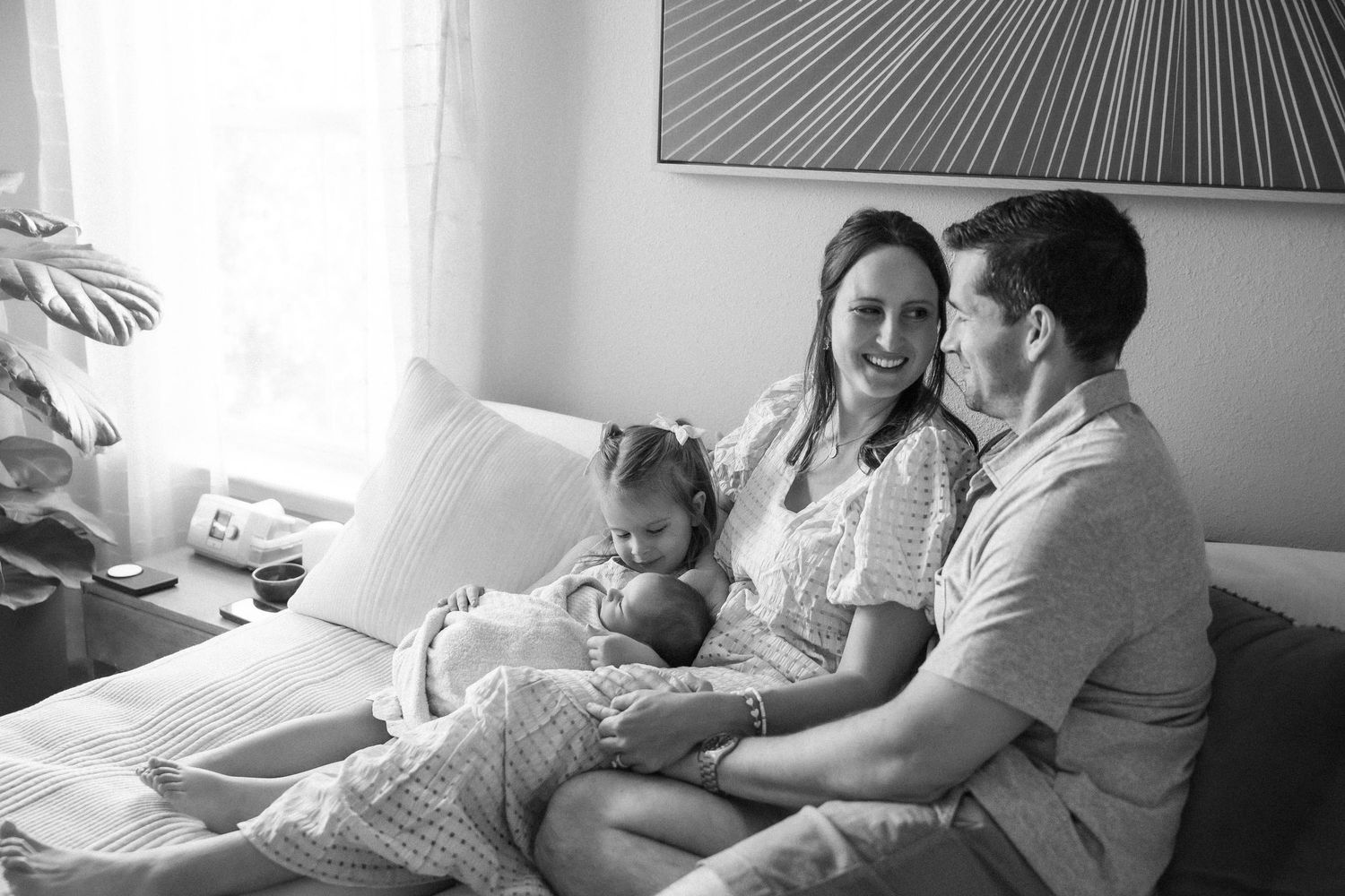 Black and white photo of a happy family with a toddler and newborn baby sitting together on a bed.
