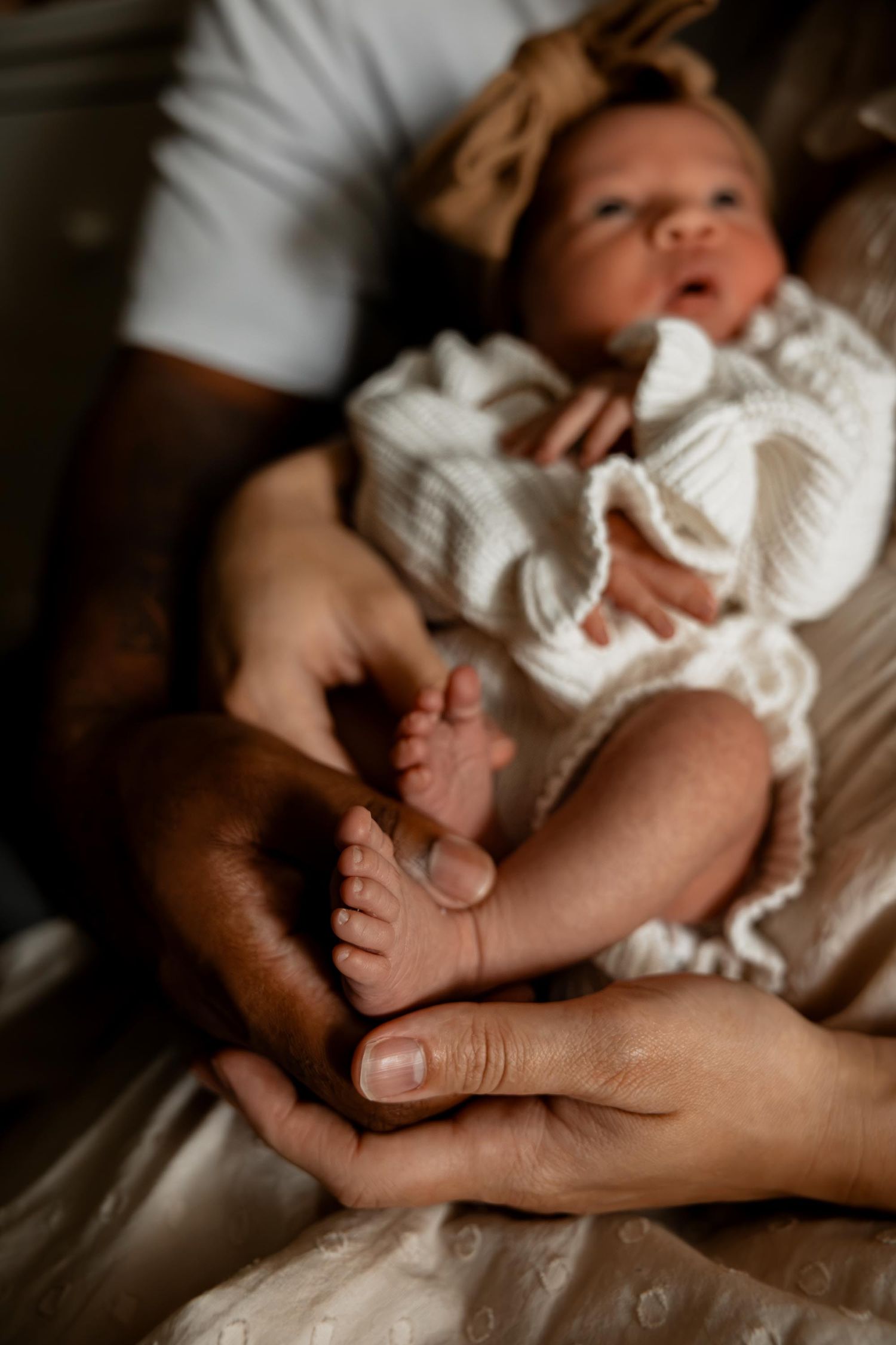 Two parents tenderly hold their newborn baby's tiny feet, baby dressed in white knit outfit.