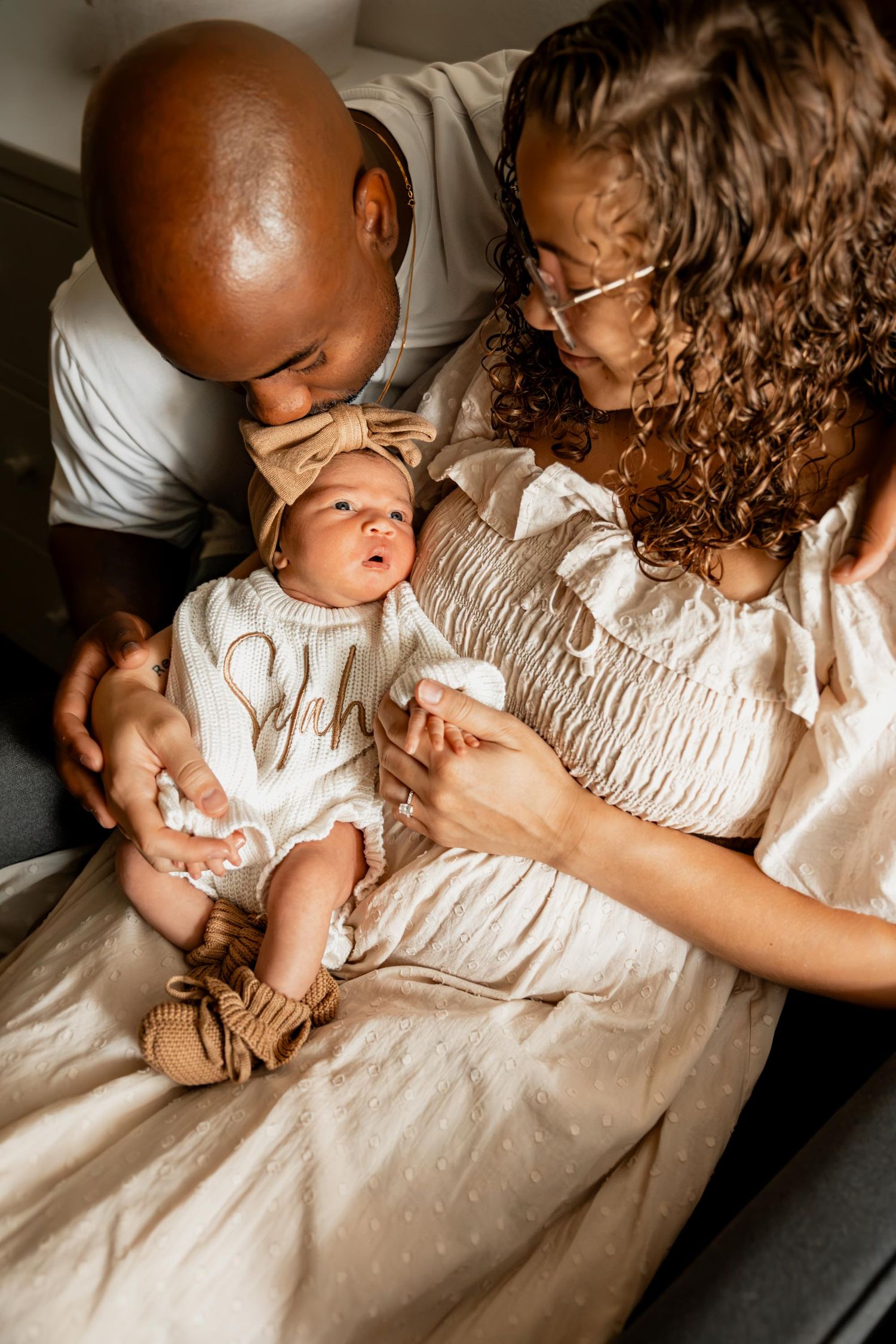 A couple lovingly gazes at their newborn baby dressed in a white outfit with a bow headband and brown sandals.