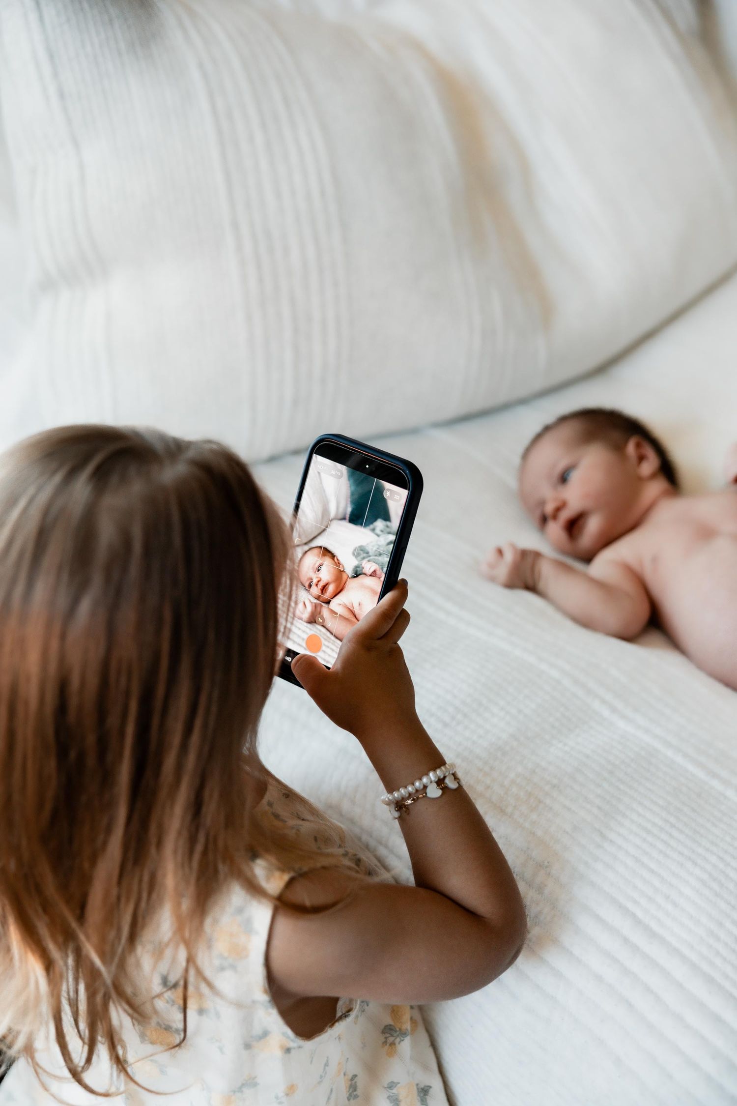 Young girl photographing a newborn baby on a white bed with her smartphone.