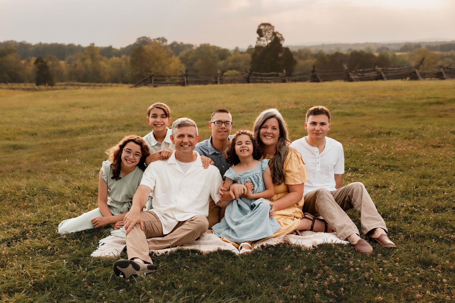 Family of seven smiling together on a blanket in a green field during golden hour outdoor photo session.